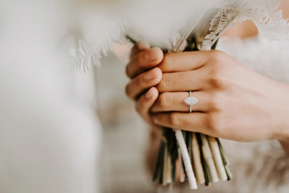 Woman holding bouquet with marquise diamond ring in white gold and traditional halo and pave set diamonds down side, bouquet is white and pale, neutral colors