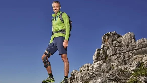 Ein Mann in Sportkleidung mit Knieorthese wandert in bergigem Gelände, trägt einen Rucksack und steht auf Felsen, vor blauem Himmel.
