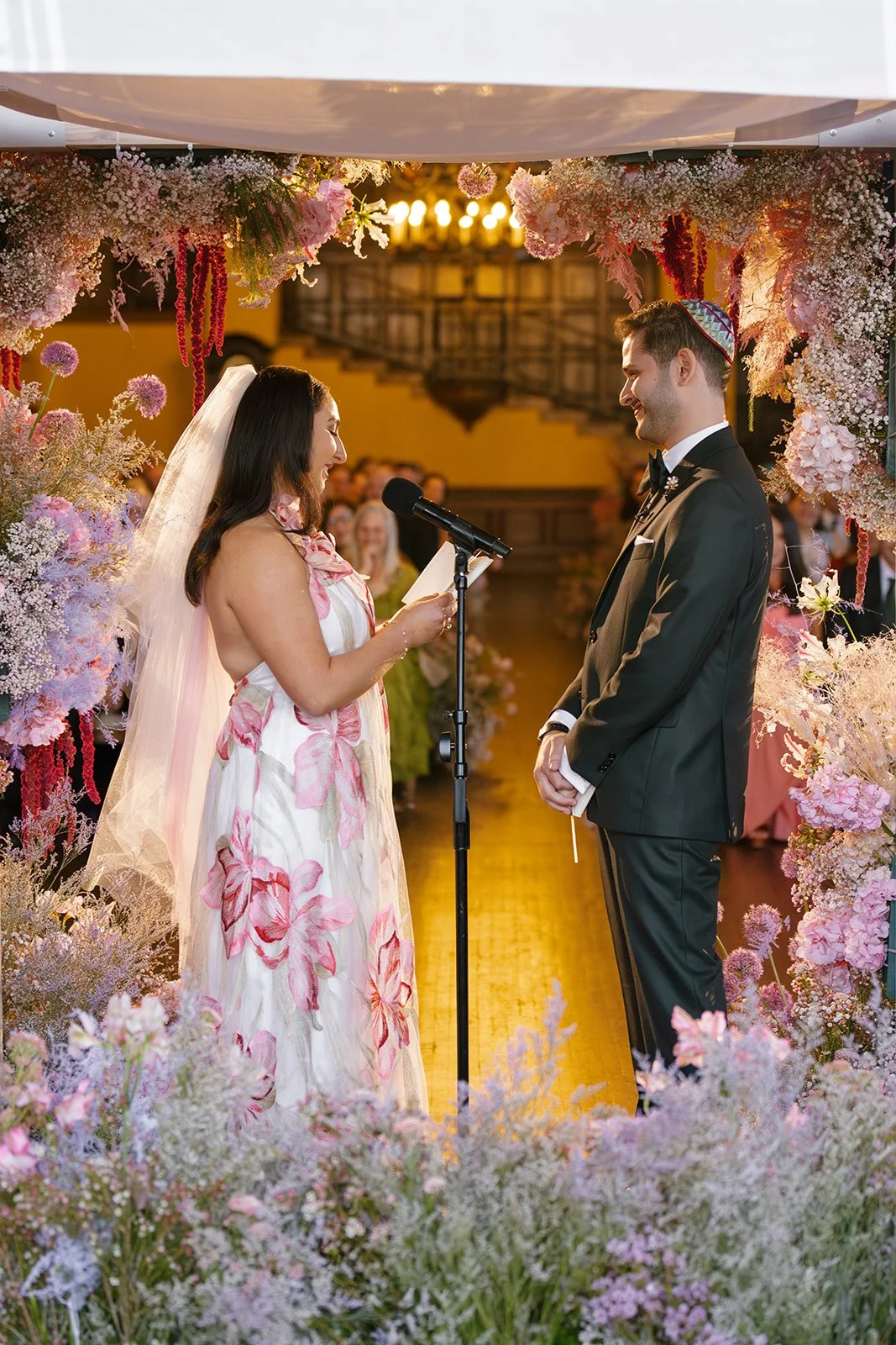 A bride in a white and pink dress with pink floral patterns and a veil reads vows into a microphone during a wedding ceremony. The groom in a black tuxedo and colorful kippah smiles at her, surrounded by pink and white flowers under a wedding chuppah