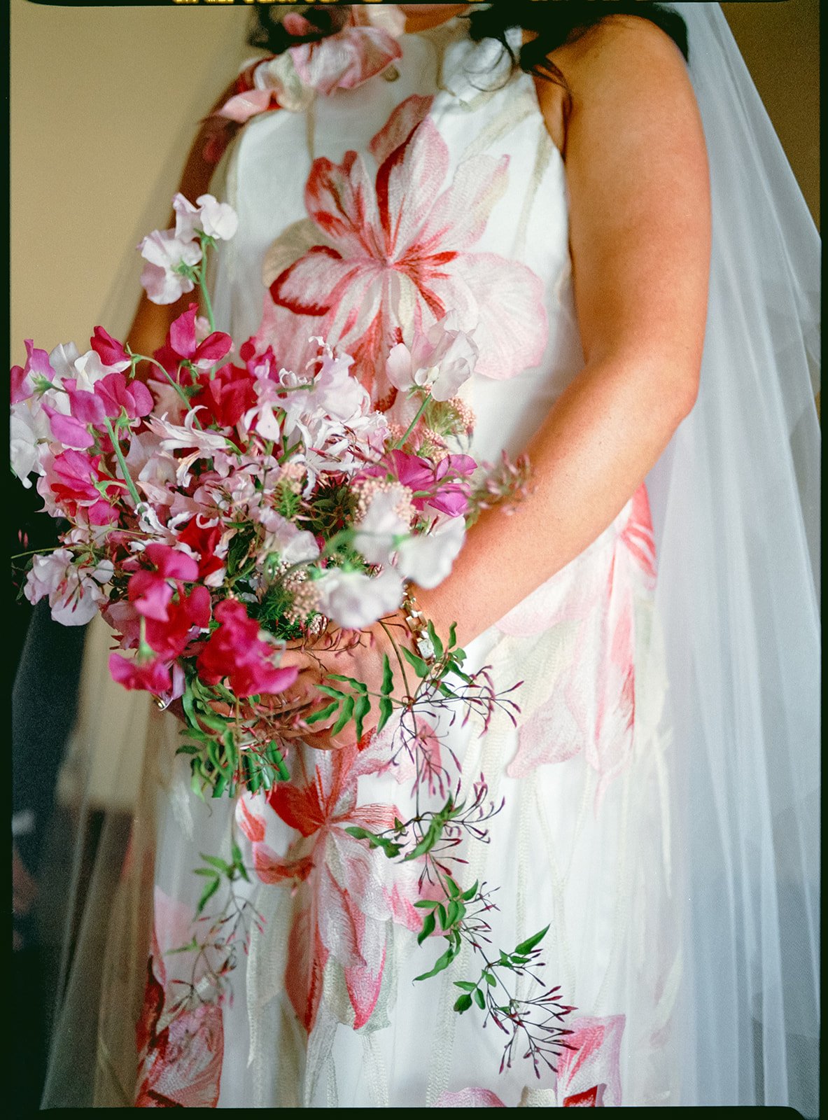 Close-up of a bride in a floral dress holding a bouquet of pink and white flowers. The dress features large pink and white floral patterns.