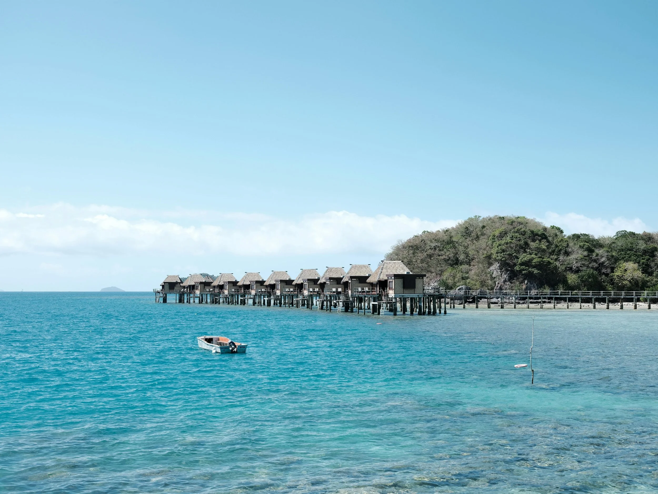 Overwater bungalows on stilts extend into clear blue ocean with a small boat floating nearby, and a green, rocky hillside in the background under a partly cloudy sky.