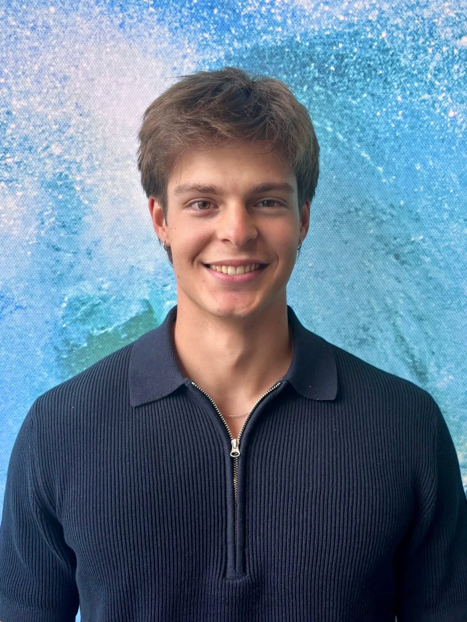 A young man with short brown hair, blue eyes, and a light complexion, smiling, wearing a dark zip-up sweater, standing in front of a sparkly blue and white abstract background.