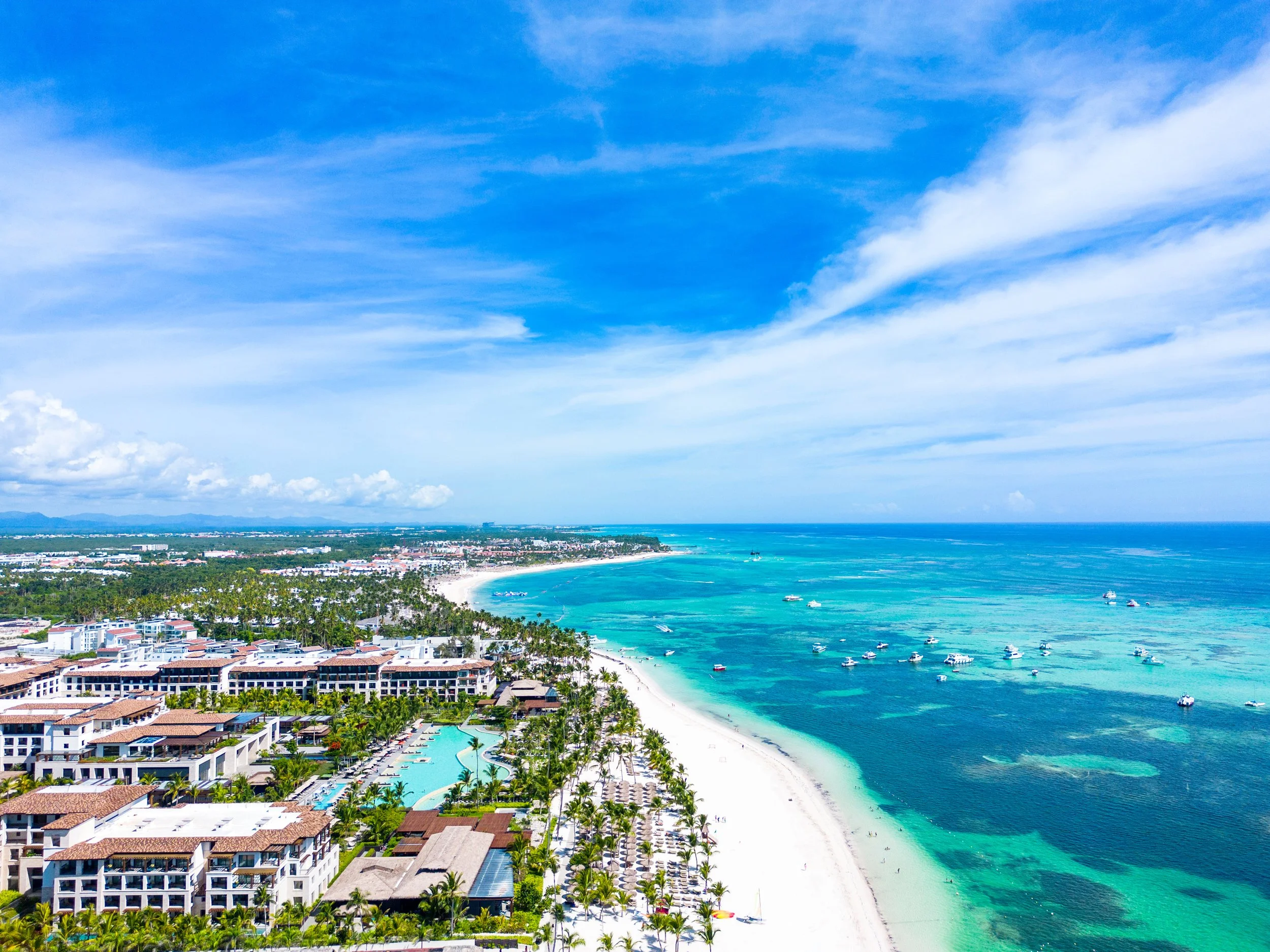 Aerial view of a resort on a tropical beach with turquoise water, palm trees, and white sand. Numerous boats are on the water and the sky is partly cloudy.