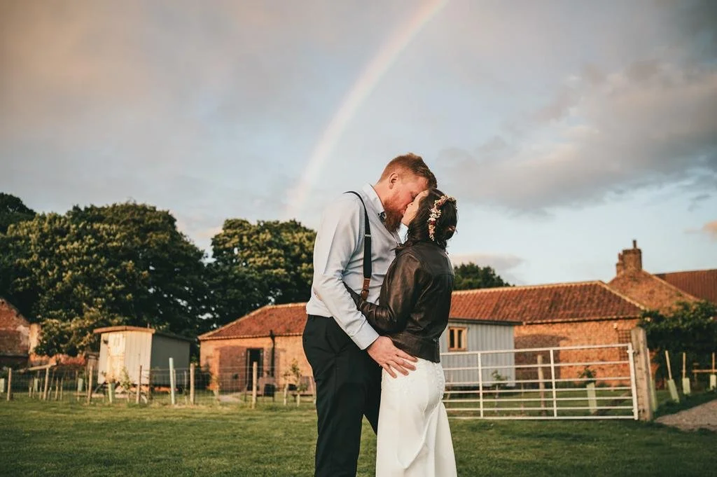 groom and bride kissing in a field with rainbow in the background