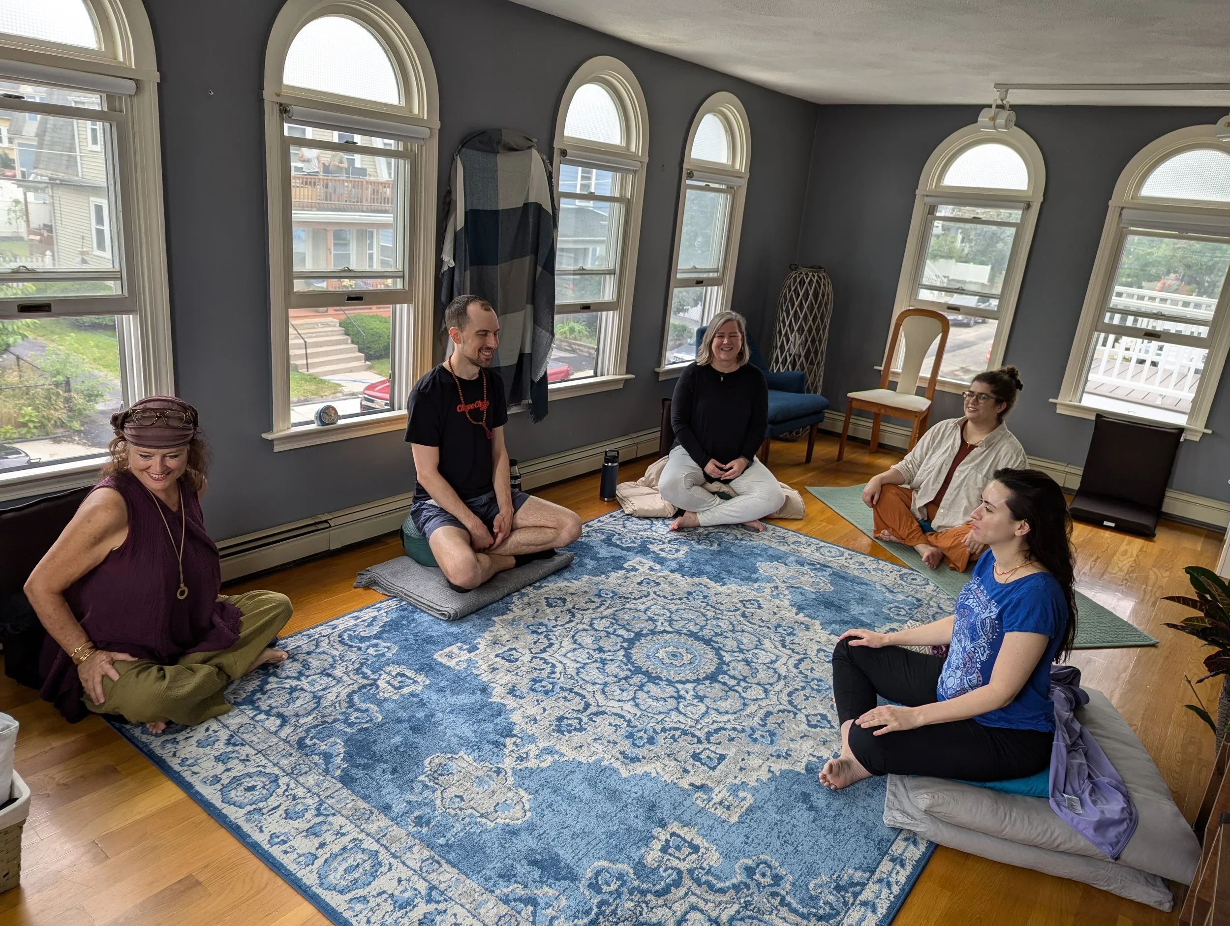 A group of six people sitting on the floor in a semi-circle, participating in a meditation or yoga session inside a room with large arched windows.