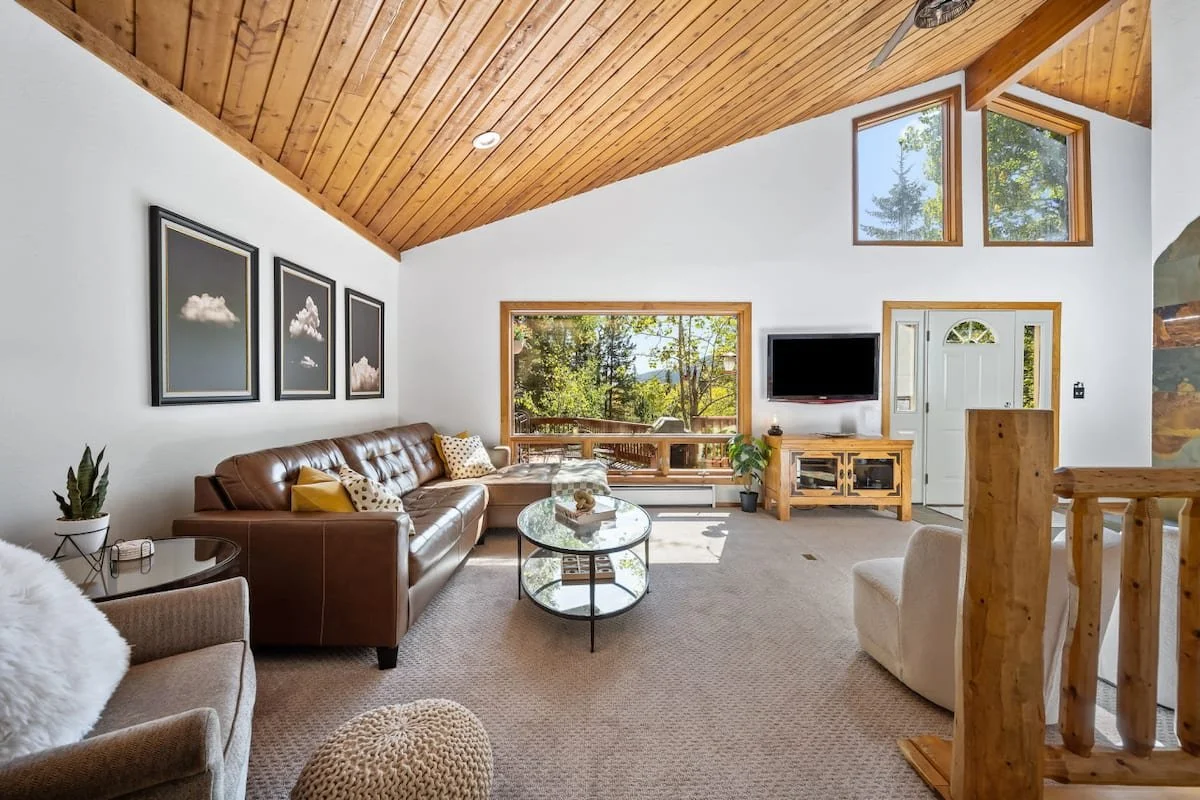 Living room with vaulted wooden ceiling, white walls, brown leather sofa, white armchair, glass coffee table, wall-mounted TV, large window showing trees outside, and wall art of clouds.
