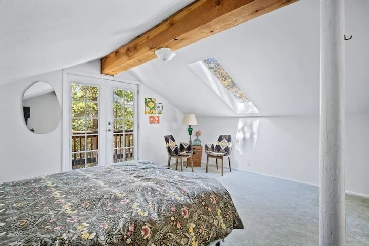 Bright bedroom with sloped ceiling, skylight window, and sliding glass doors leading to a balcony, with a bed, two chairs, and a small side table.