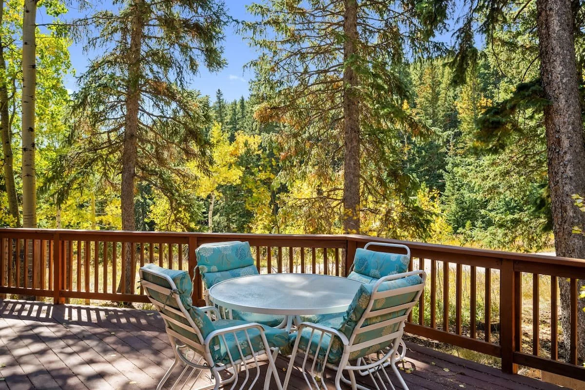 A wooden deck with a round table and cushioned chairs surrounded by tall trees and lush foliage.