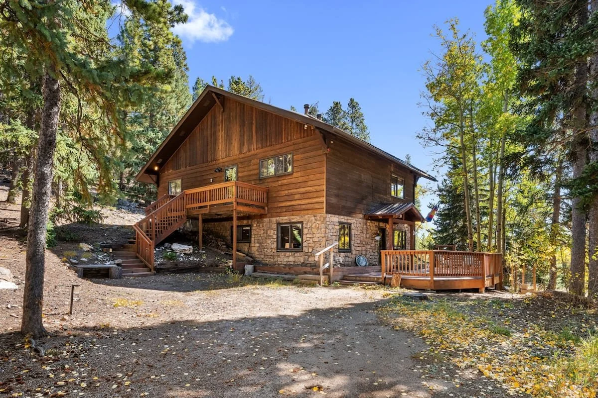 A wooden house in a forested area with trees surrounding it, a stone foundation, and multiple decks, some with railings, on a sunny day.