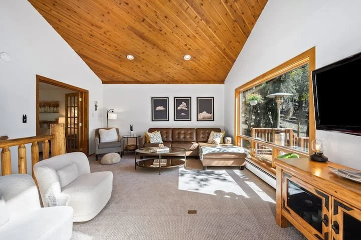 Living room with a wooden vaulted ceiling, white walls, a large window, a sectional sofa with pillows, framed wall art, a white armchair, a wooden entertainment console, and a flat-screen TV.