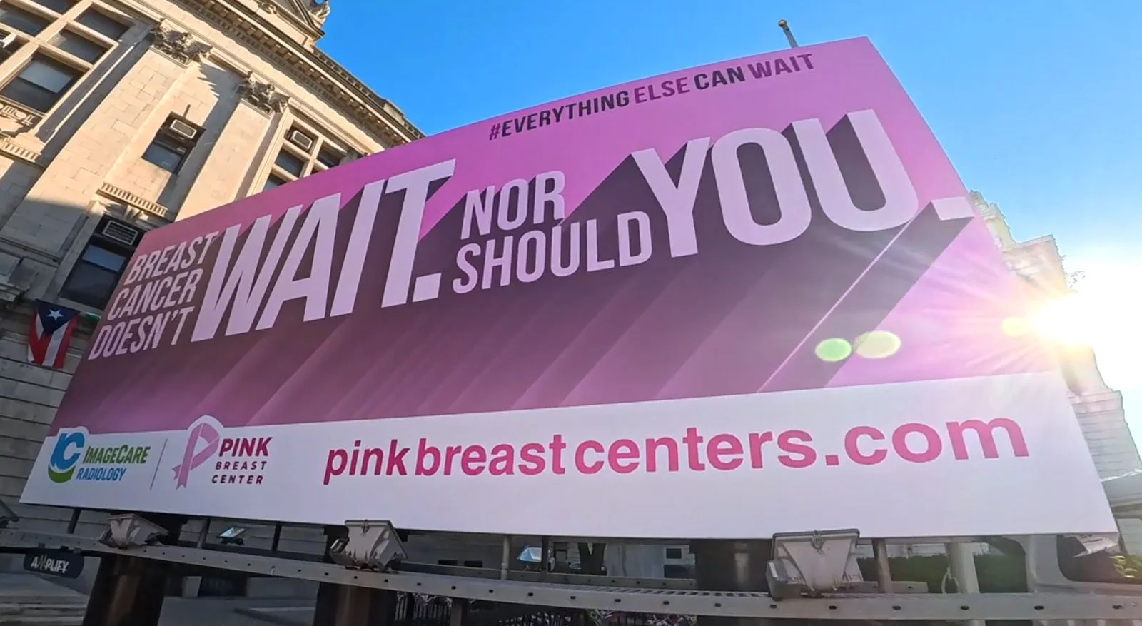 Large pink billboard with the message that breast cancer doesn't wait, nor should you, promoting breast cancer awareness. The billboard includes logos for IMAGECARE Radiology and PINK Breast Center, and the website pinkbreastcenters.com, with a city building and sunny sky in the background.