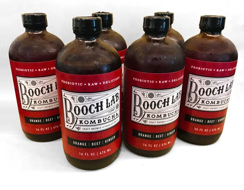 Six bottles of Booch Lab Kombucha with orange, beet, and ginger flavor on a plain white surface.