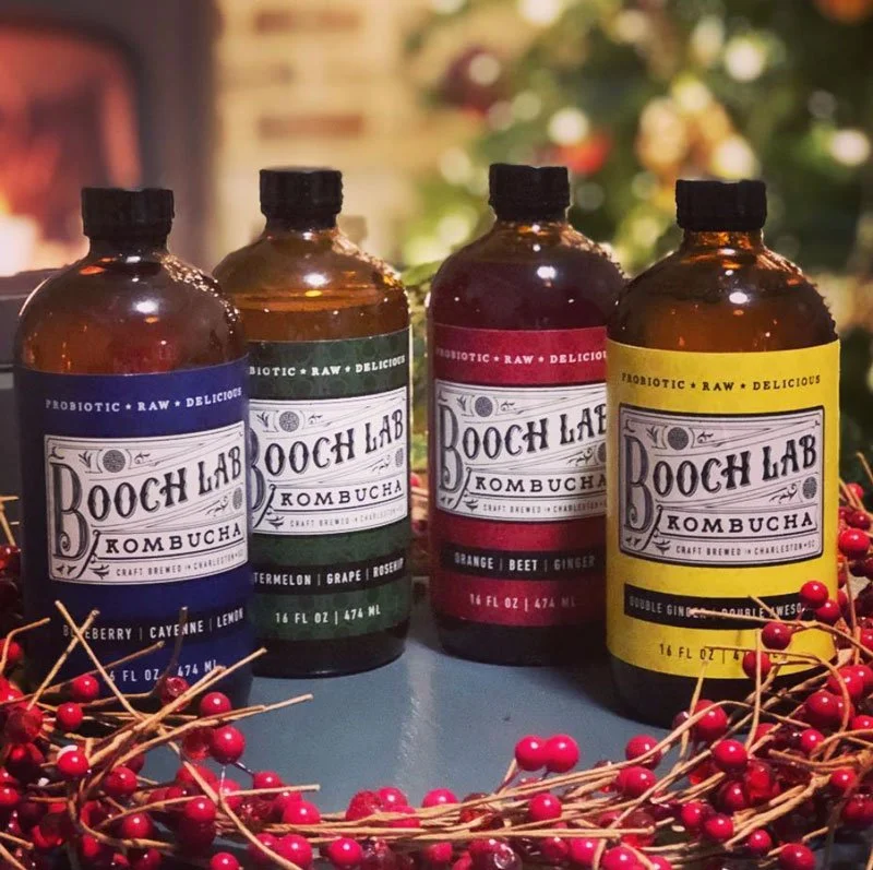 Four bottles of Booch Lab Kombucha in blue, green, red, and yellow labels, surrounded by red berries on branches, with a blurred Christmas tree in the background.