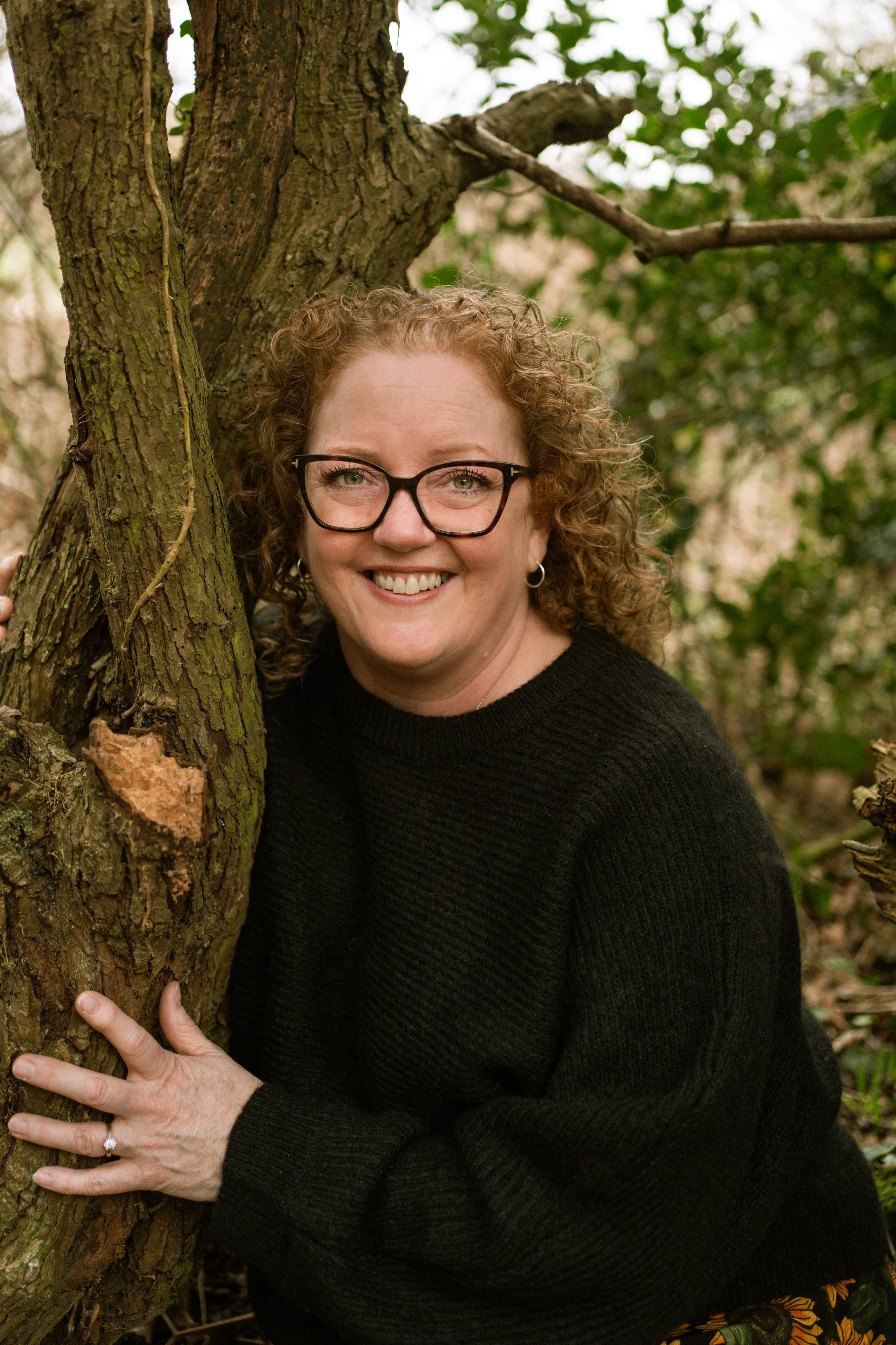 A woman with curly hair and glasses smiling, leaning against a tree in a forest, wearing a black sweater and floral skirt.