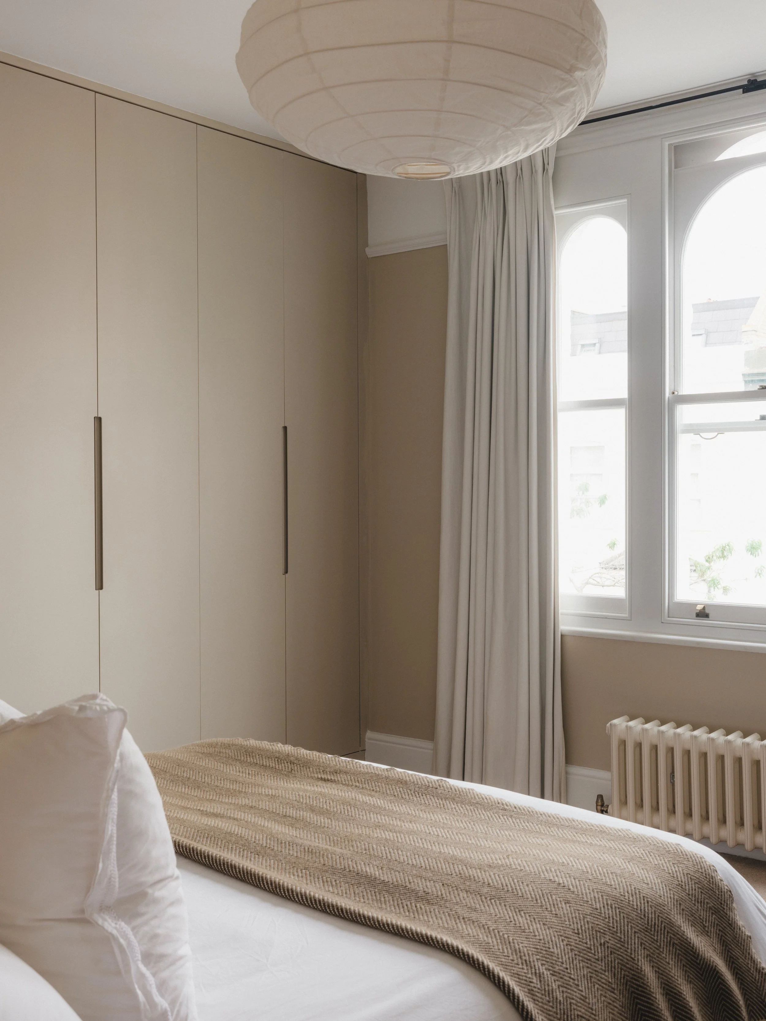 Bedroom with beige walls, large pendant light, built-in wardrobe, bed with white linens and a brown blanket, and window with white curtains.