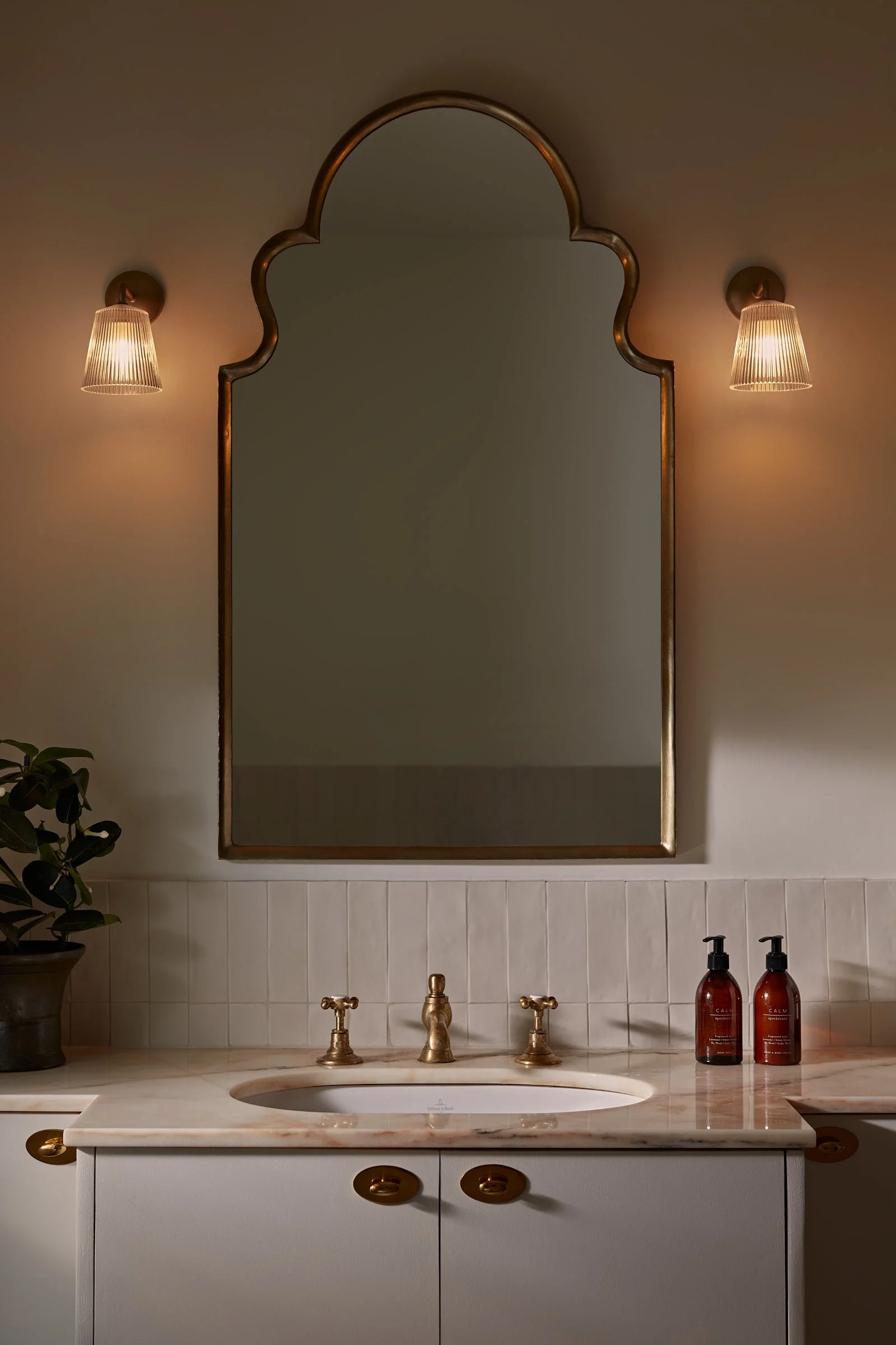 Bathroom vanity with marble top, brass taps, arched mirror and wall lights in a warm, layered scheme, designed by Partner London