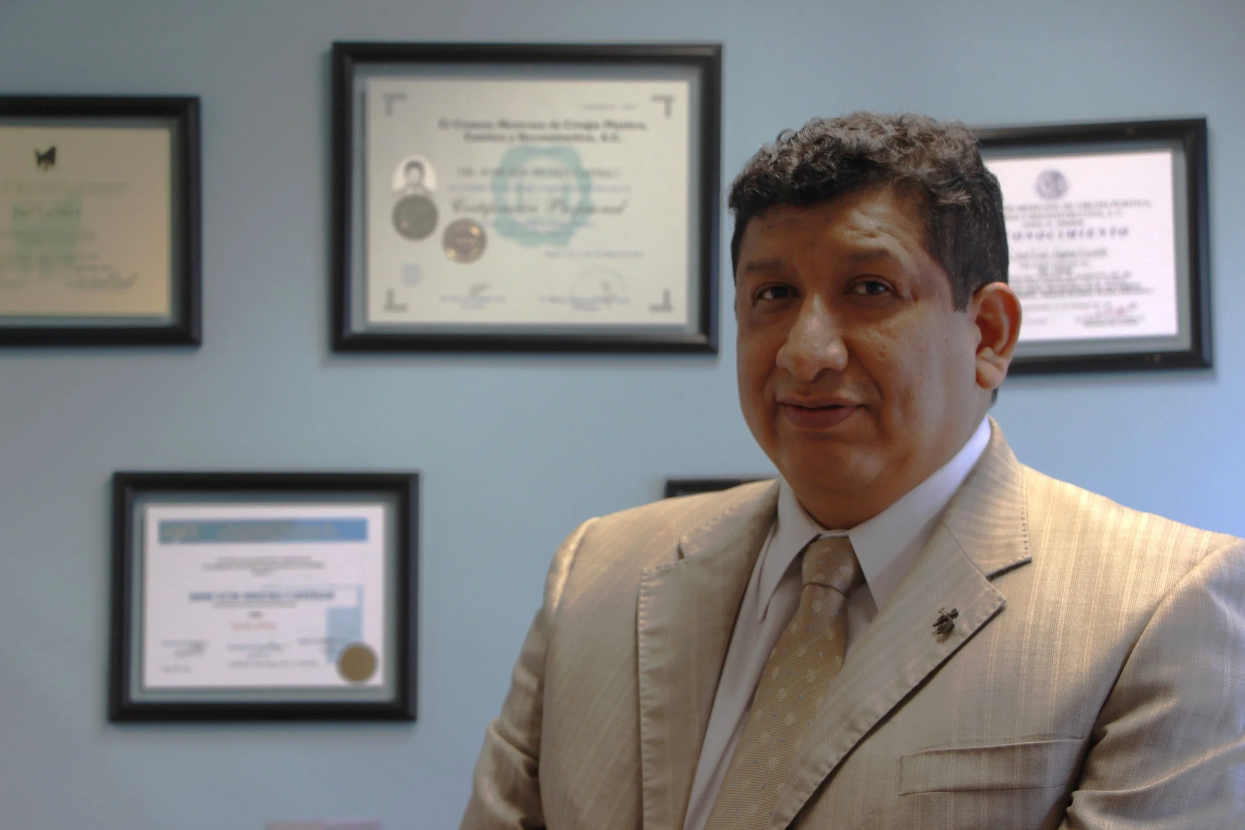 Hombre con traje beige en oficina con certificados y diplomas enmarcados en la pared.