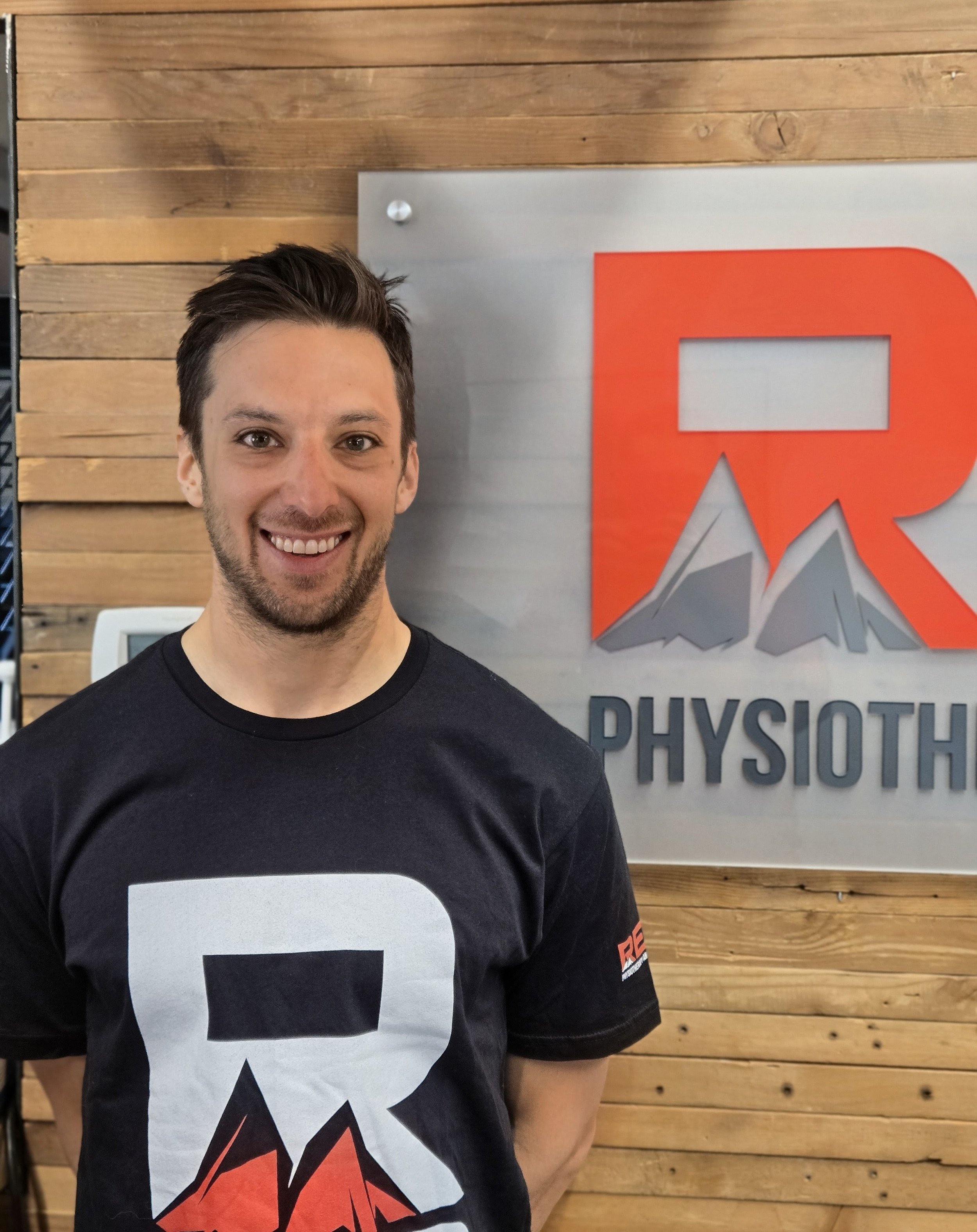 Man smiling in front of a sign that reads 'PHYSIOTHERAPY' with a mountain graphic and a wooden wall background.