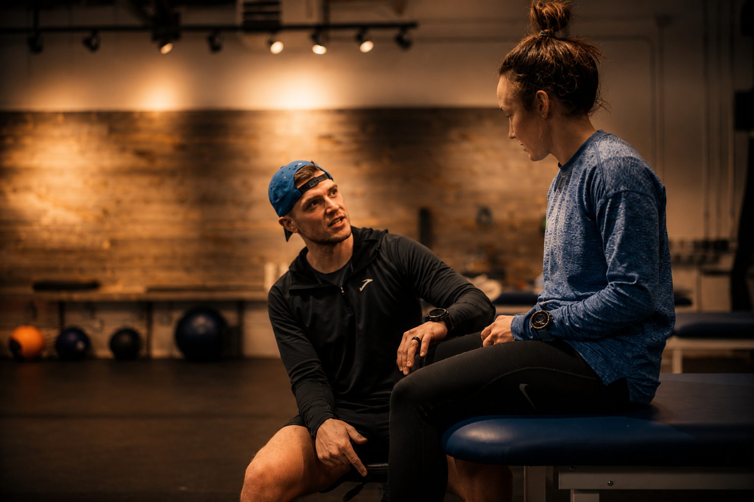 A man and a woman in workout attire having a conversation in a gym, with the man sitting on a bench and the woman standing and leaning forward.