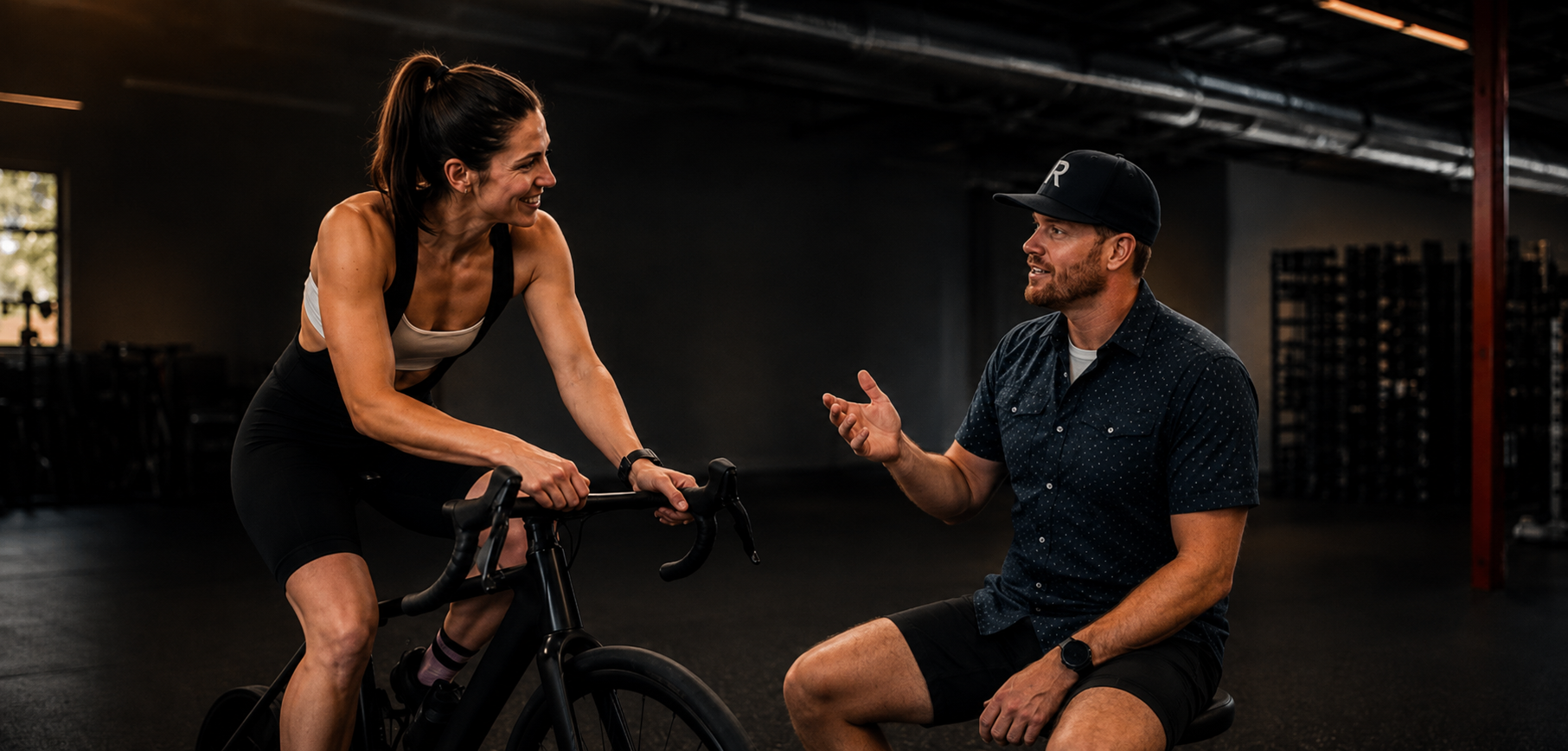 A woman on a bicycle having a conversation with a man sitting nearby in a gym.