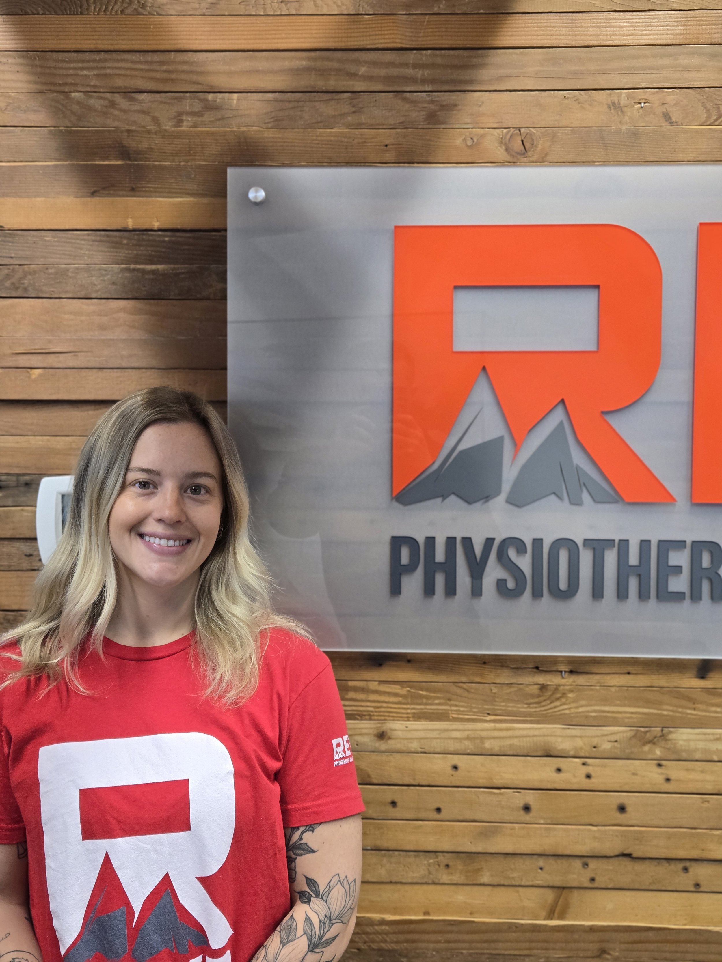 A woman with blonde hair smiling, wearing a red t-shirt, standing in front of a wooden wall and a sign that says 'PHYSIOTHERAPY'. Only part of her face and upper body are visible in the image.