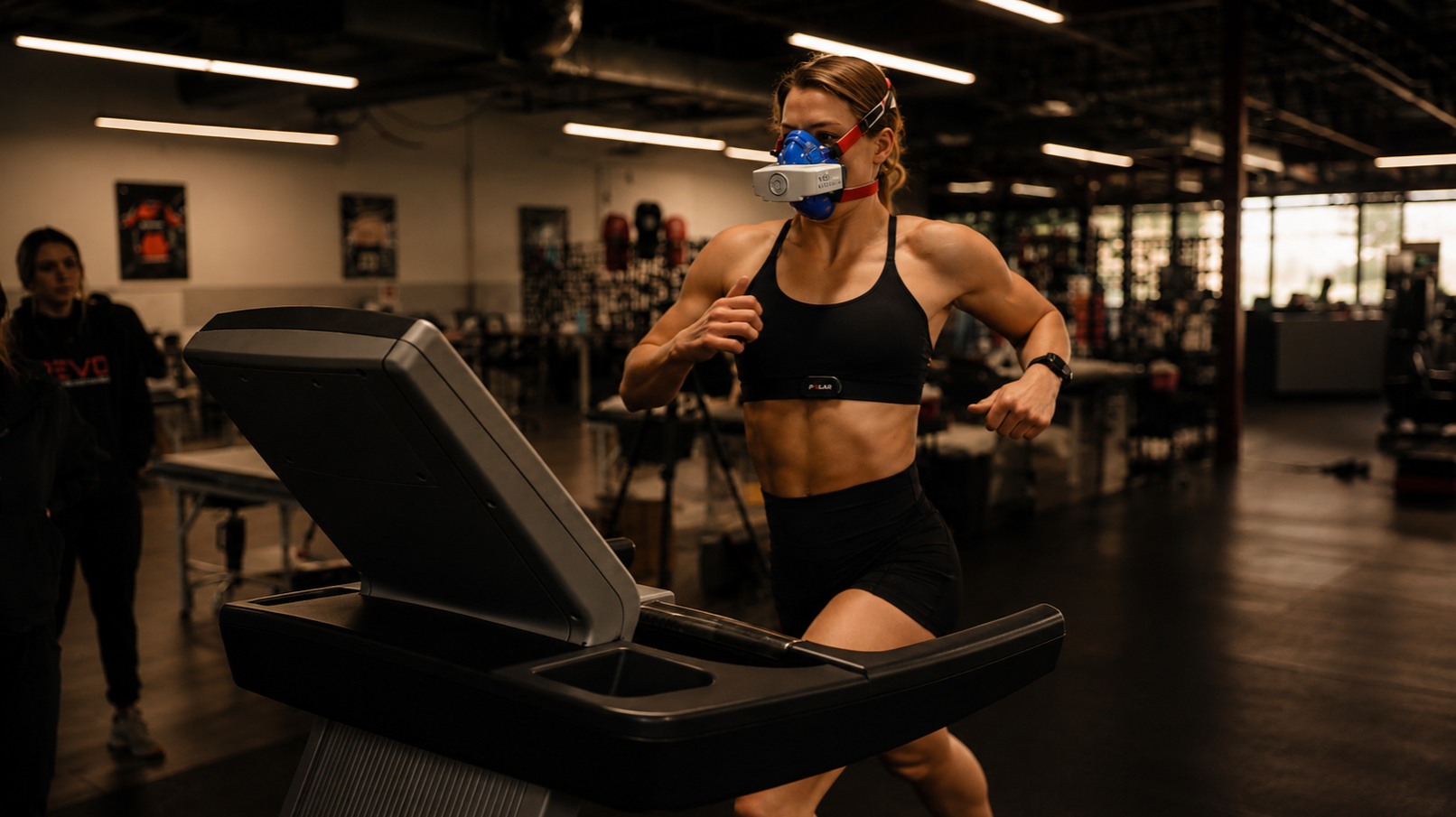 A woman with an athletic build running on a treadmill in a gym, wearing a black sports bra, black shorts, a blue face mask with a head strap, and a fitness tracker on her wrist. There are two women watching her in the background.