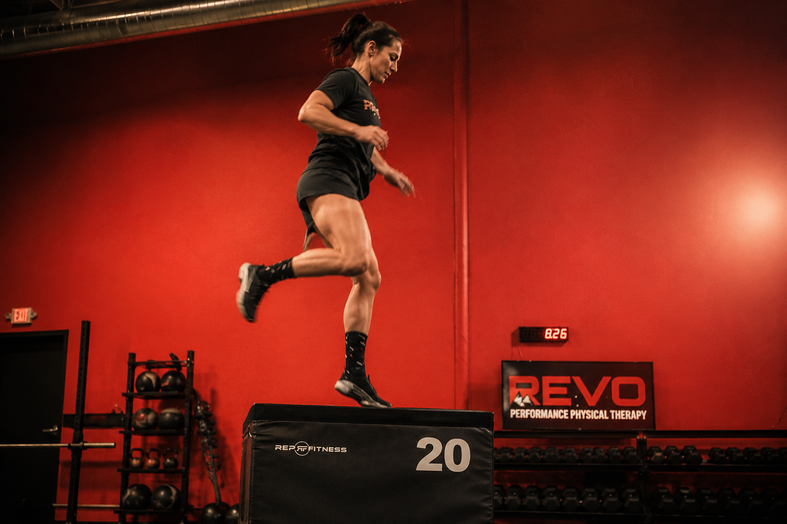 A woman leaps onto a black box in a gym with red walls, black shelves with kettlebells, and a sign that reads "REVO Performance Physical Therapy."