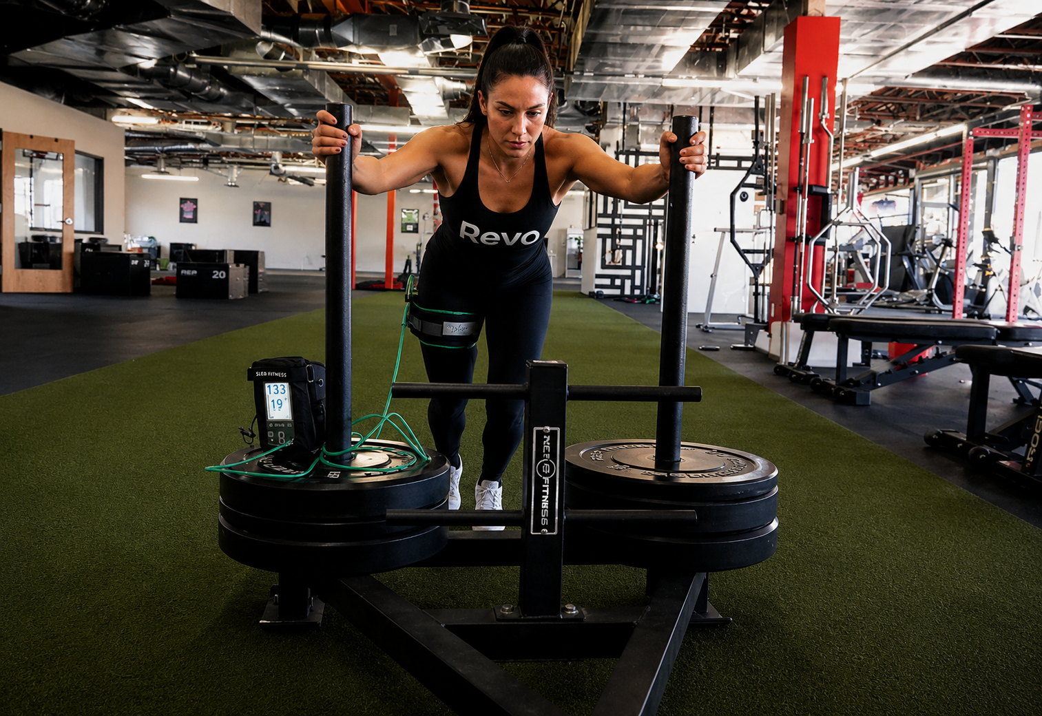 A woman working out on a sled push machine at a gym, wearing black workout clothes and a knee brace, with gym equipment visible in the background.