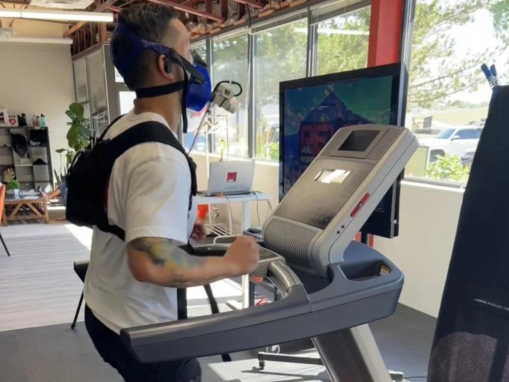 Person running on a treadmill in an indoor gym, wearing headphones and a face mask, with a large screen in front displaying a scenic mountain landscape and windows letting in natural light.