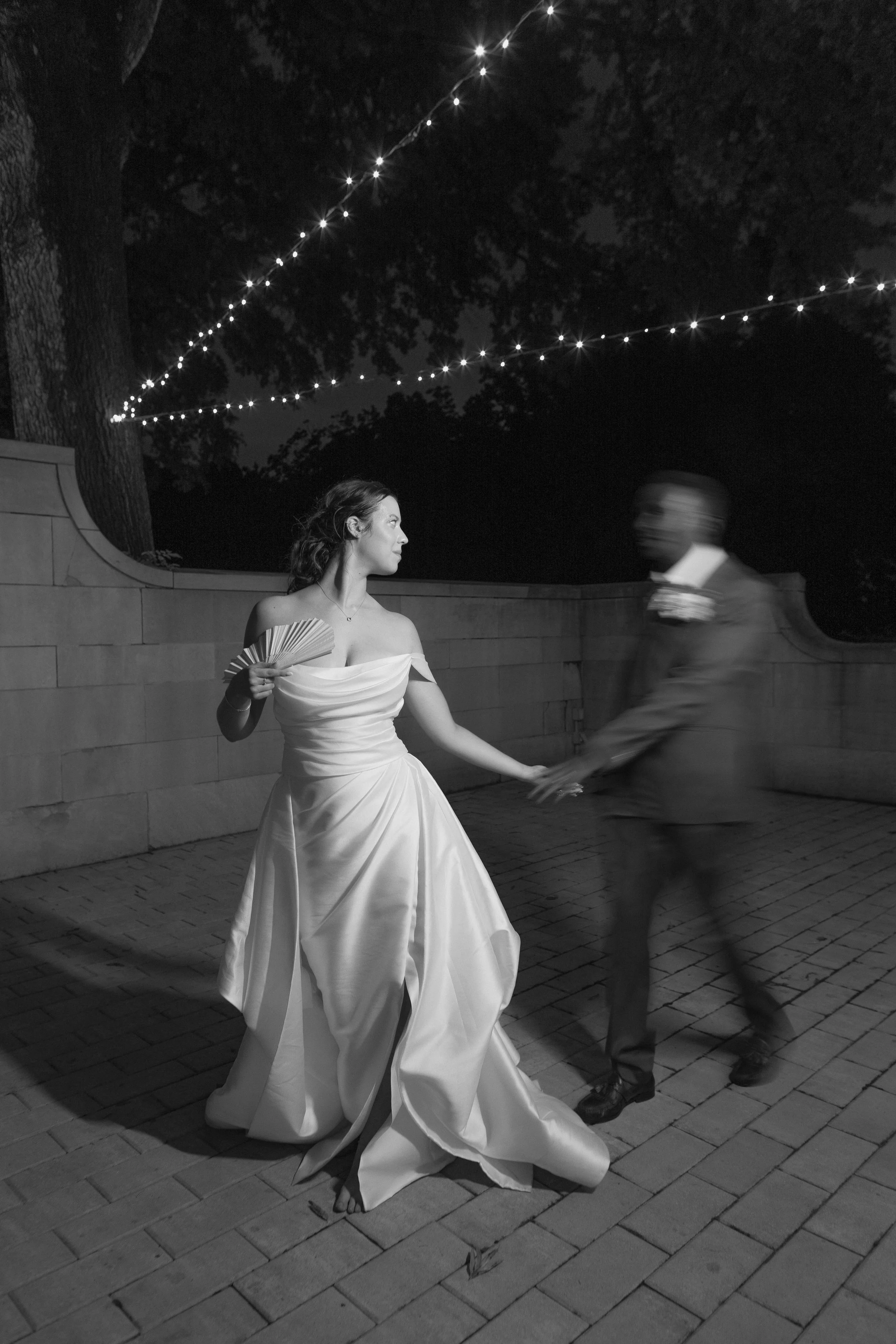 Bride twirling with the groom under string lights, black and white, Jillian and César's wedding at Curtis Arboretum