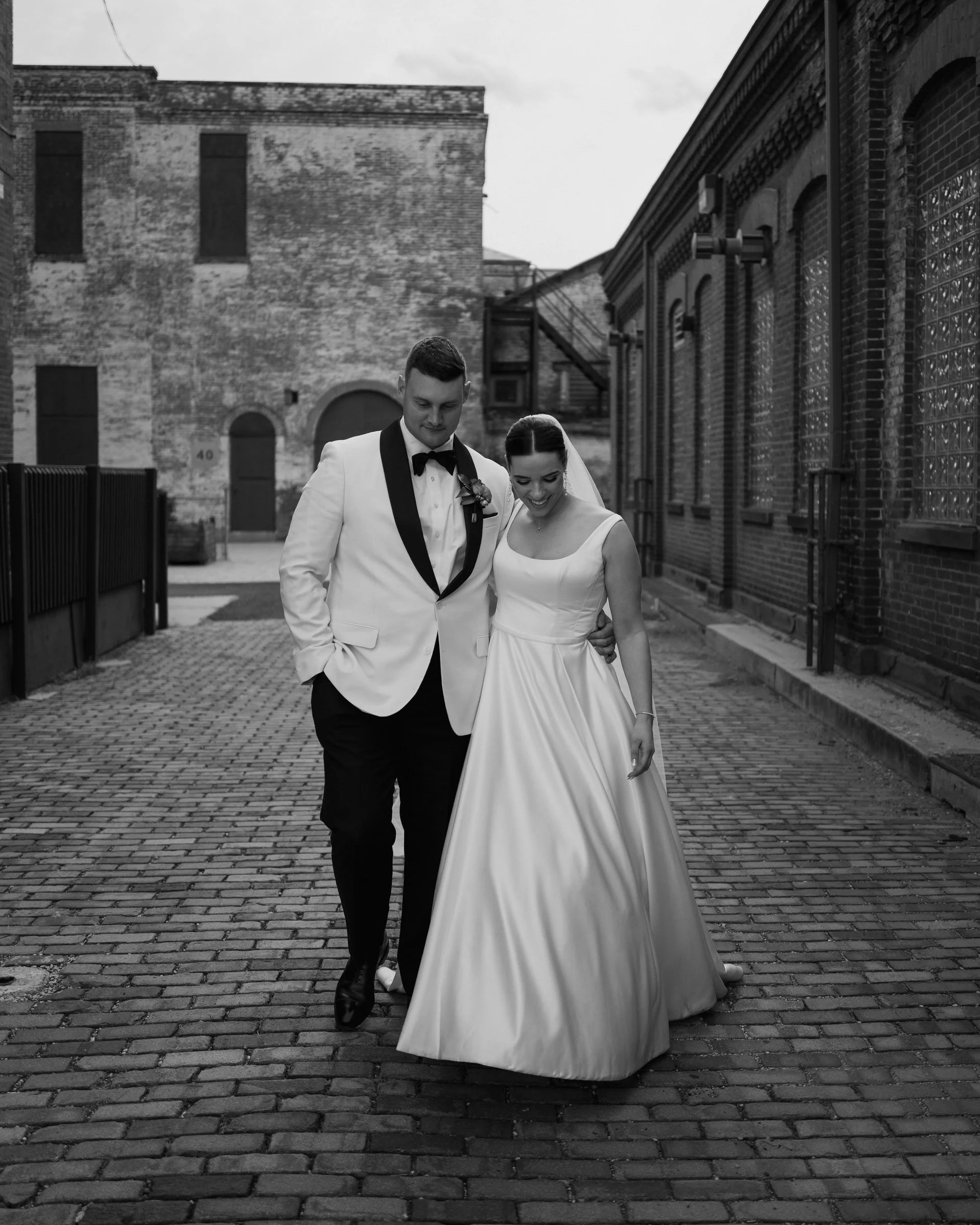 A bride and groom walking together on a cobblestone street, both smiling and looking down, with the bride wearing a long satin wedding dress and the groom in a tuxedo with a bow tie. The background features brick and stone buildings, and the photo is in black and white.