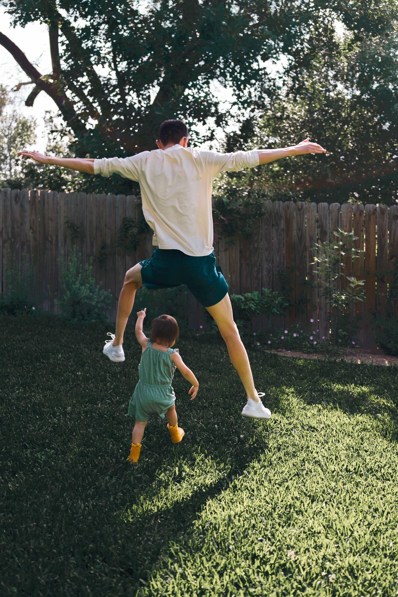 A young man is jumping in a backyard with his arms and legs extended, while a small girl in a green dress and yellow boots watches. The scene is outdoors on a grassy lawn with trees and a wooden fence in the background.