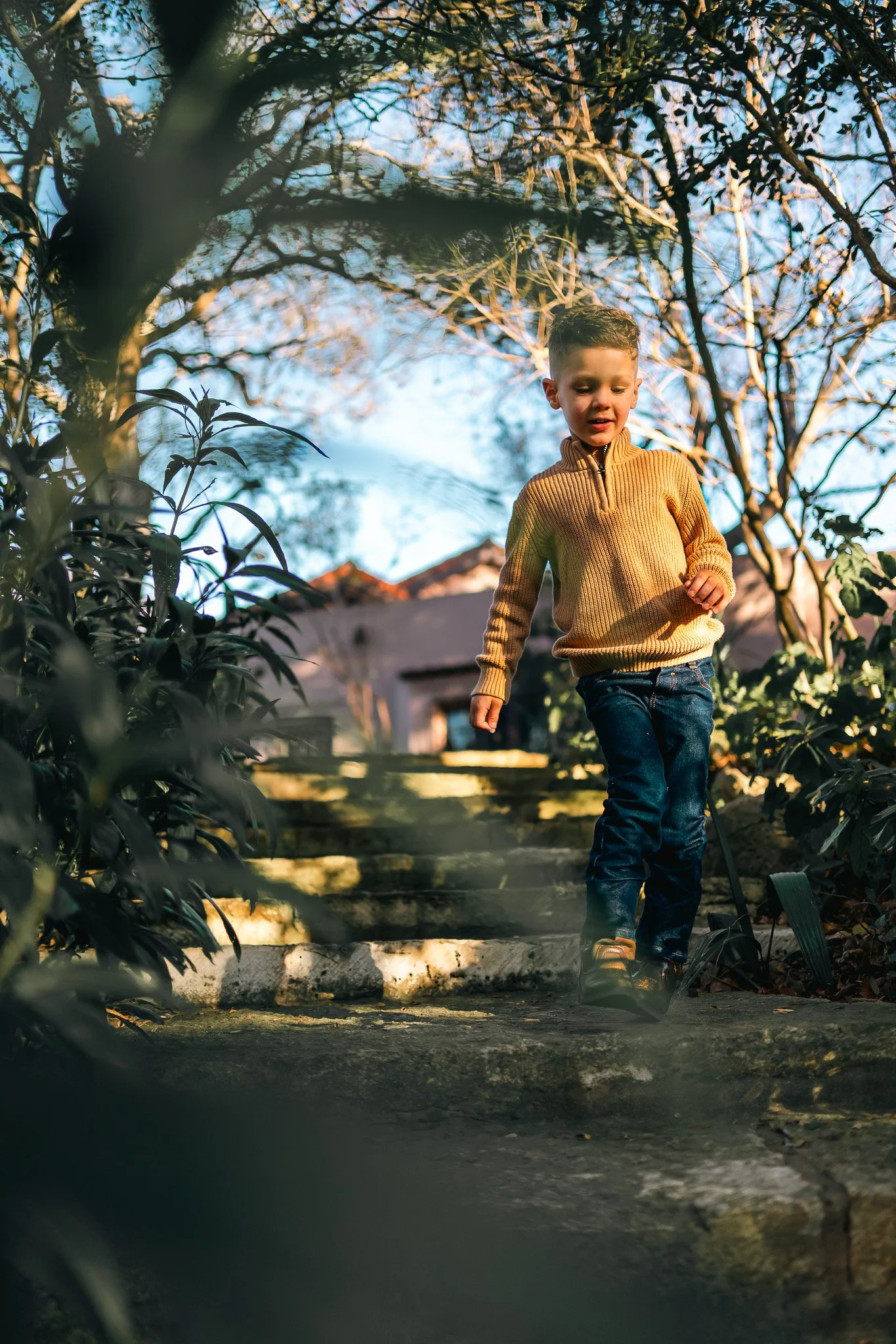 A young boy in a mustard yellow sweater and blue jeans walking down outdoor stone stairs surrounded by greenery and trees, with a house in the background on a sunny day.