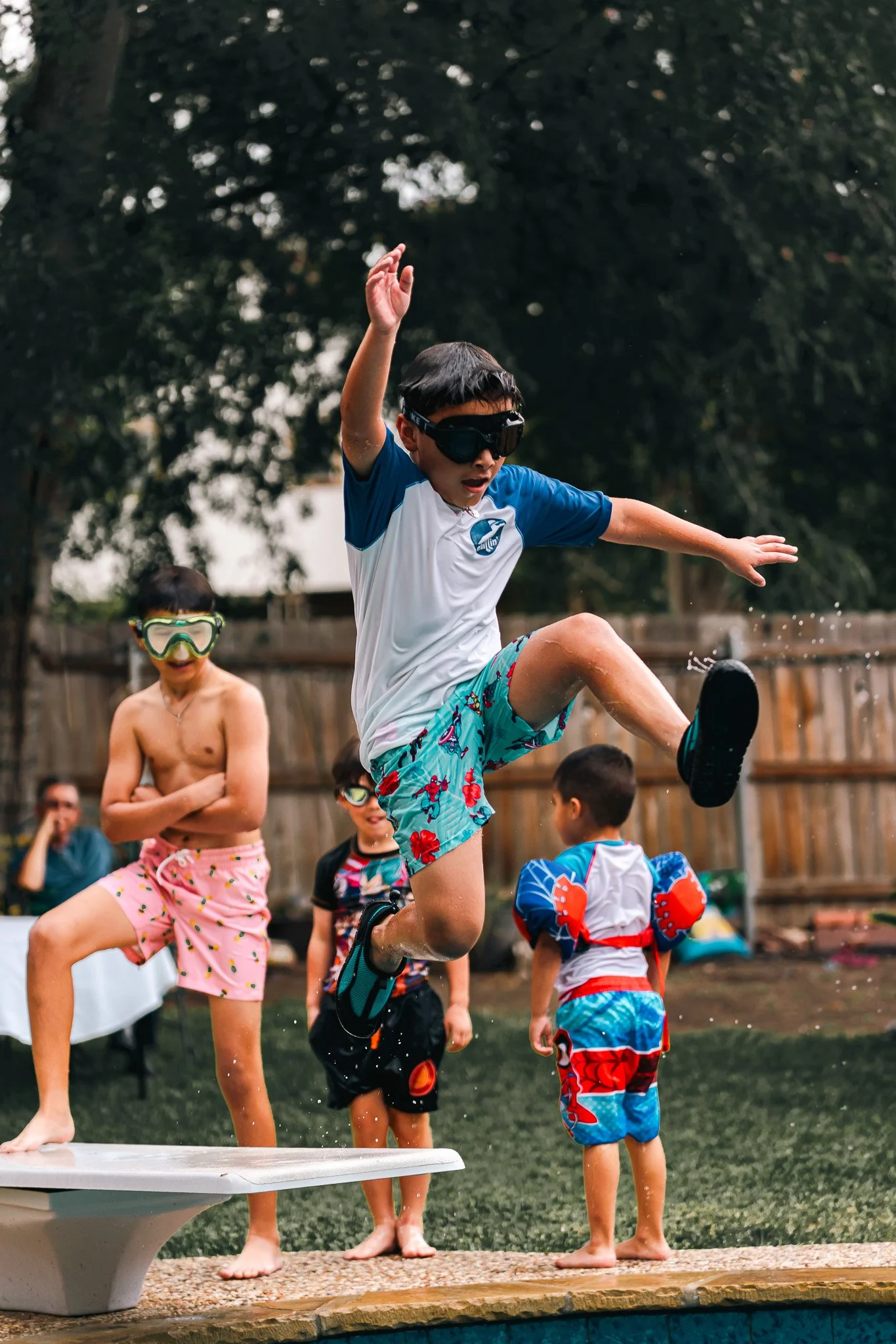 Children playing and jumping into a backyard swimming pool, wearing swimsuits and goggles, with a fence and trees in the background.