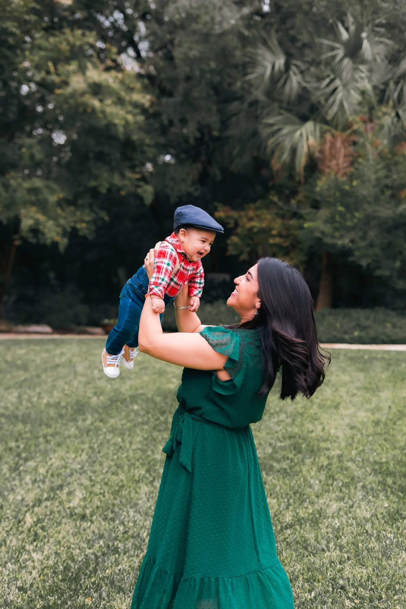 A woman in a green dress joyfully holding a young boy in a red plaid shirt and jeans, playing in a park with trees in the background.