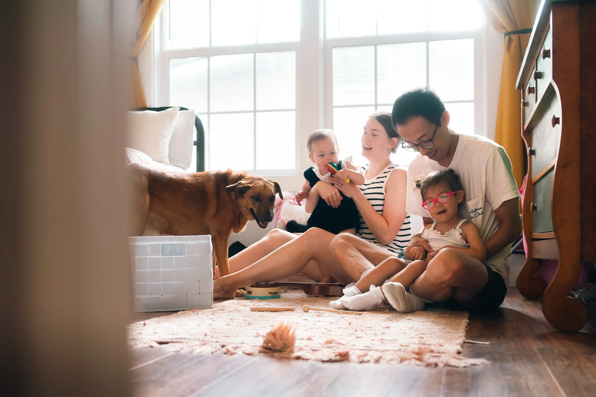A family of four with a dog sitting on the floor in a sunlit bedroom, playing with toys and laughing.