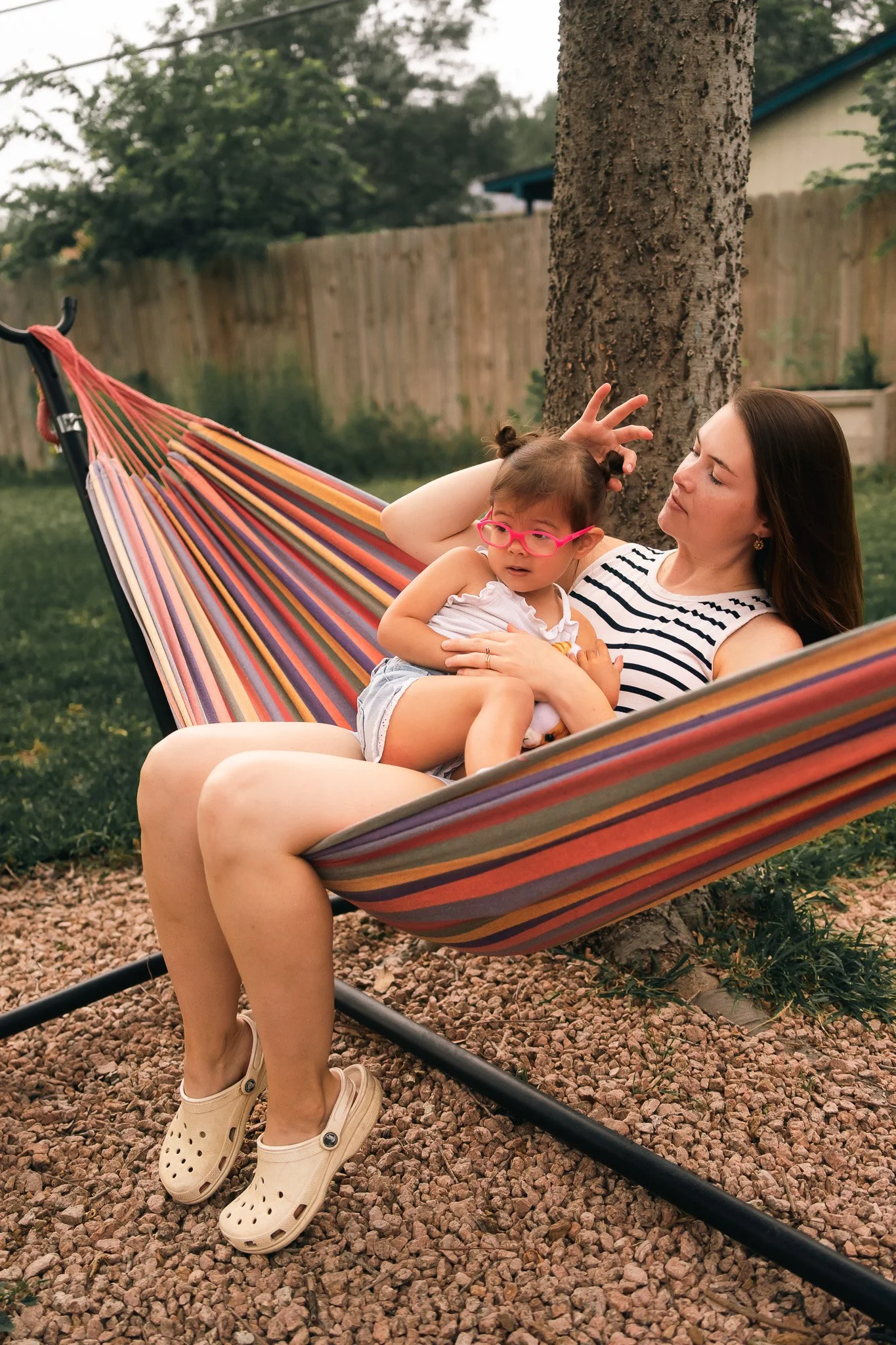 A woman lounging in a colorful hammock outdoors, holding a young girl with glasses and a white shirt, both leaning against a tree.