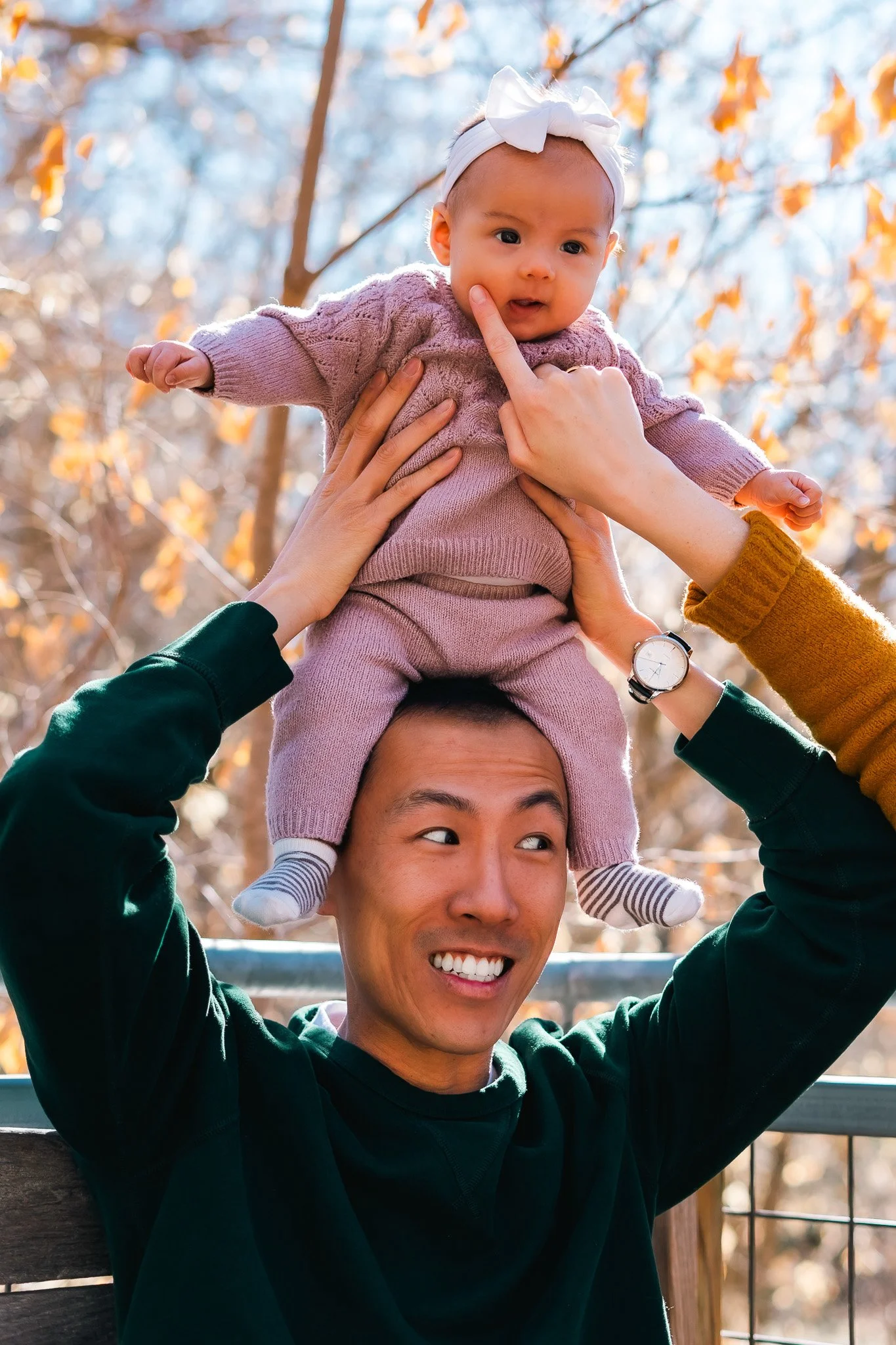 A man is holding a baby girl on his shoulders outdoors during fall, with trees and autumn leaves in the background. The baby is wearing a pink outfit and a white bow, and appears curious or surprised.