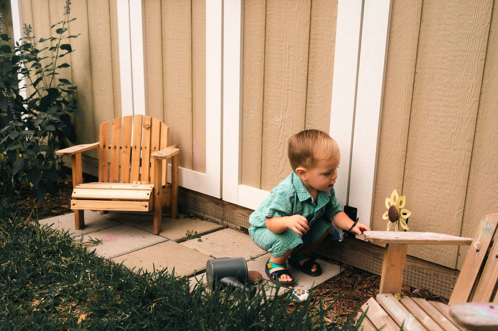 A young boy crouching outdoors next to a wooden fence, holding a small decorative sunflower, with wooden chairs and greenery nearby.