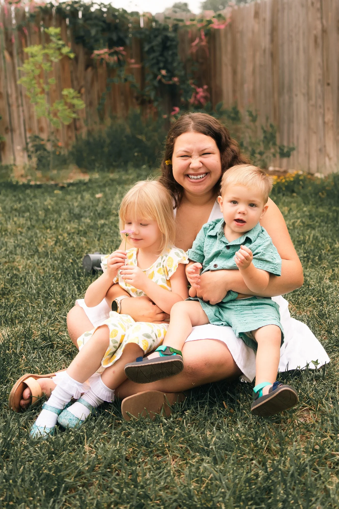 A smiling woman sitting on grass with two young children, one girl and one boy, in a backyard with a wooden fence and greenery.
