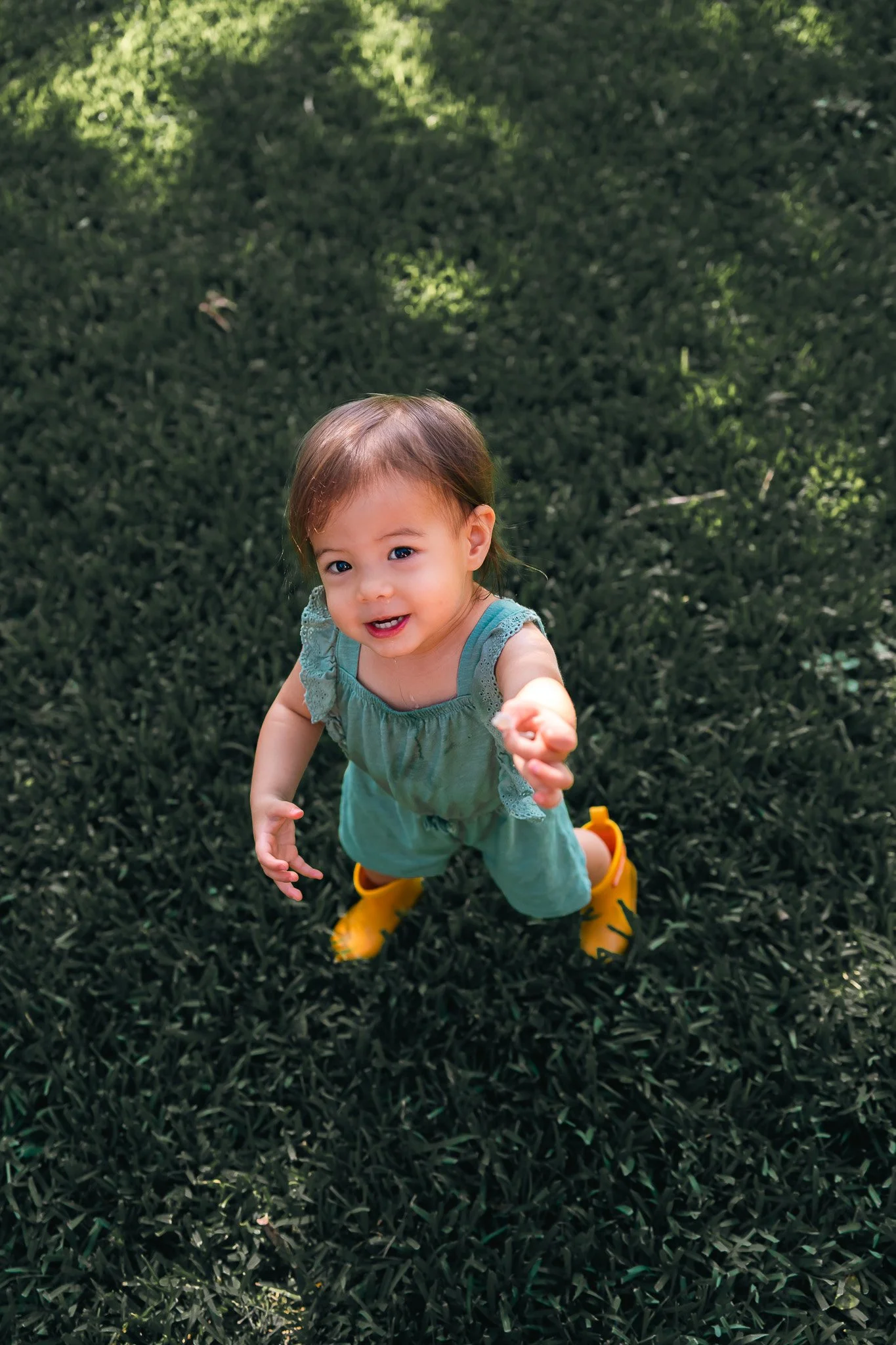 A young girl with brown hair, wearing a turquoise dress and yellow rain boots, standing on grass and looking up with a smile.