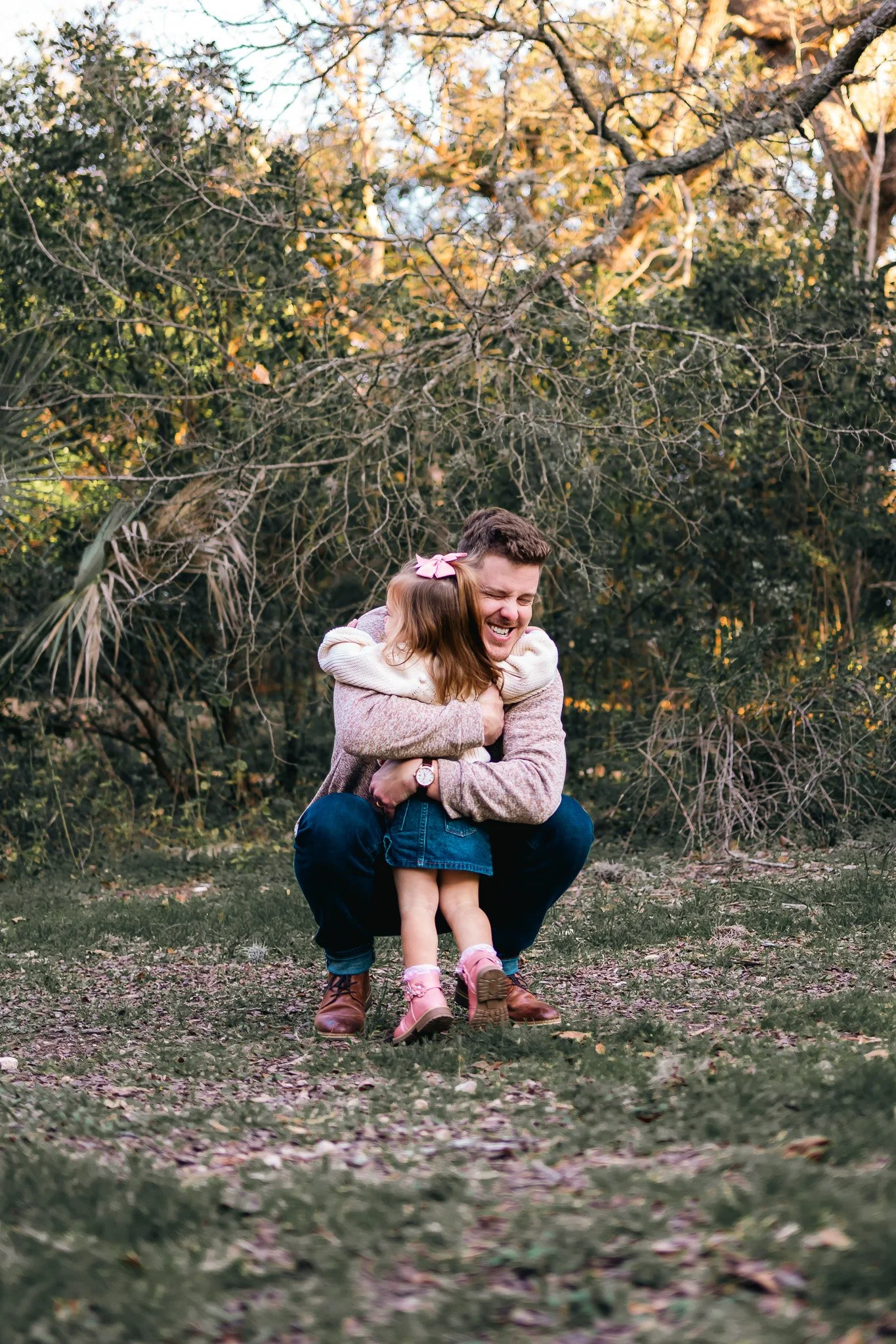A man and young girl hugging outdoors in a wooded area, both smiling joyfully.