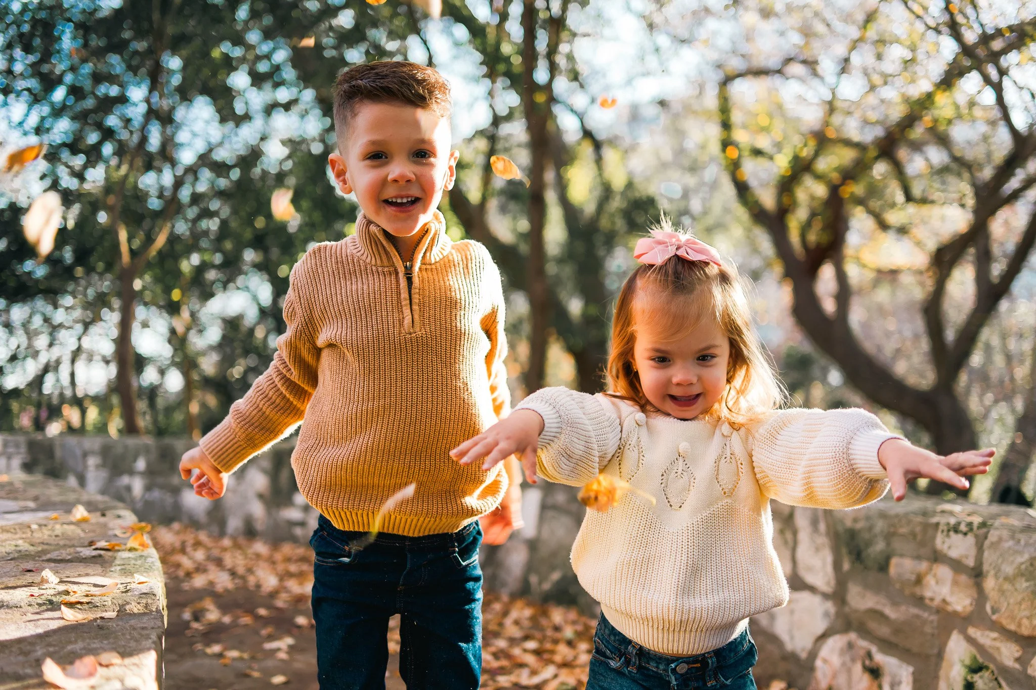 Two children, a boy and a girl, playing outdoors in a park during fall with leaves falling around them. The boy is wearing a yellow sweater and the girl is wearing a white sweater with a pink bow in her hair.