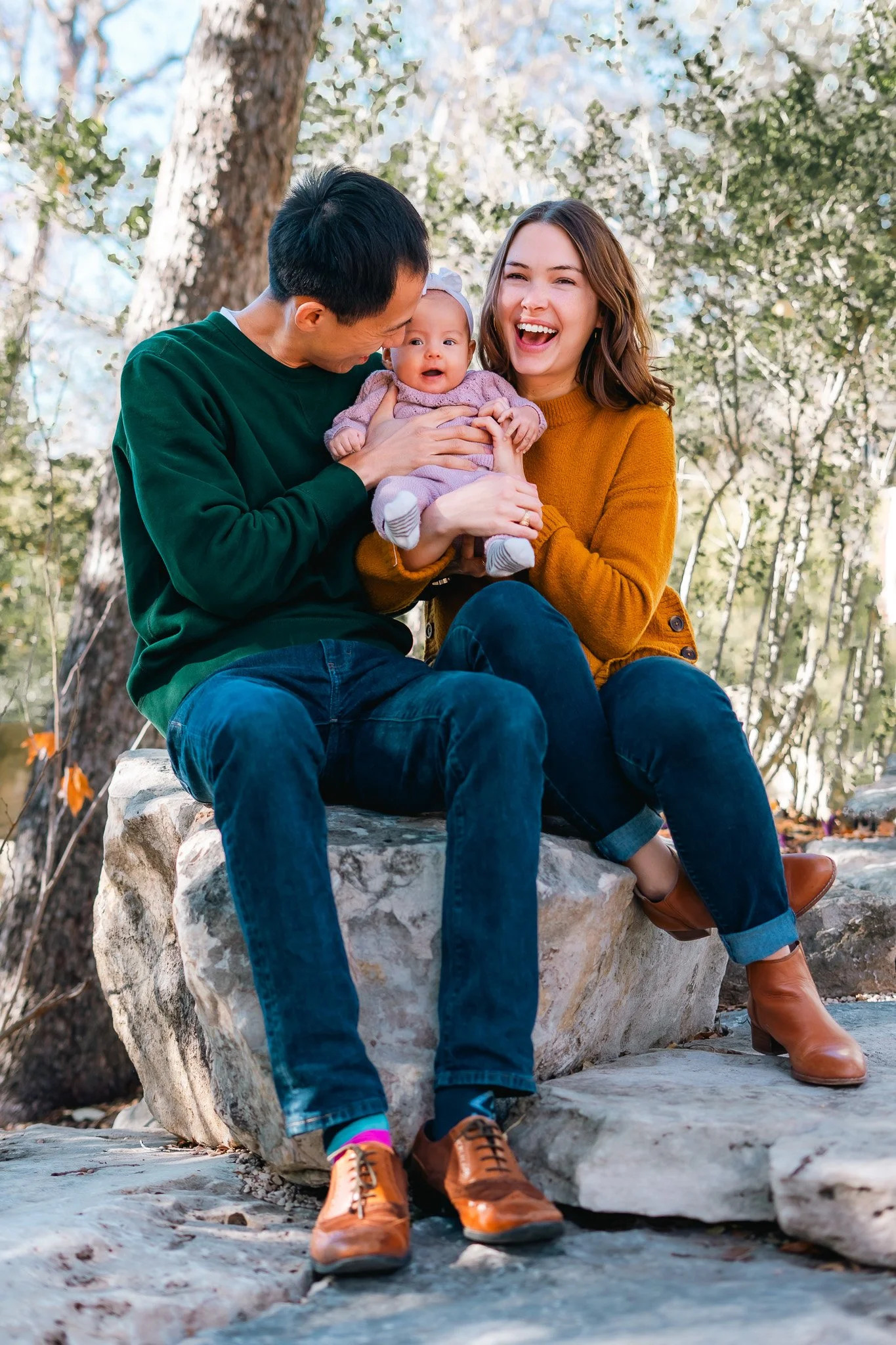 A family of three sitting on a large rock outdoors in a wooded area, smiling and enjoying time together. The father, mother, and their baby girl are dressed casually.