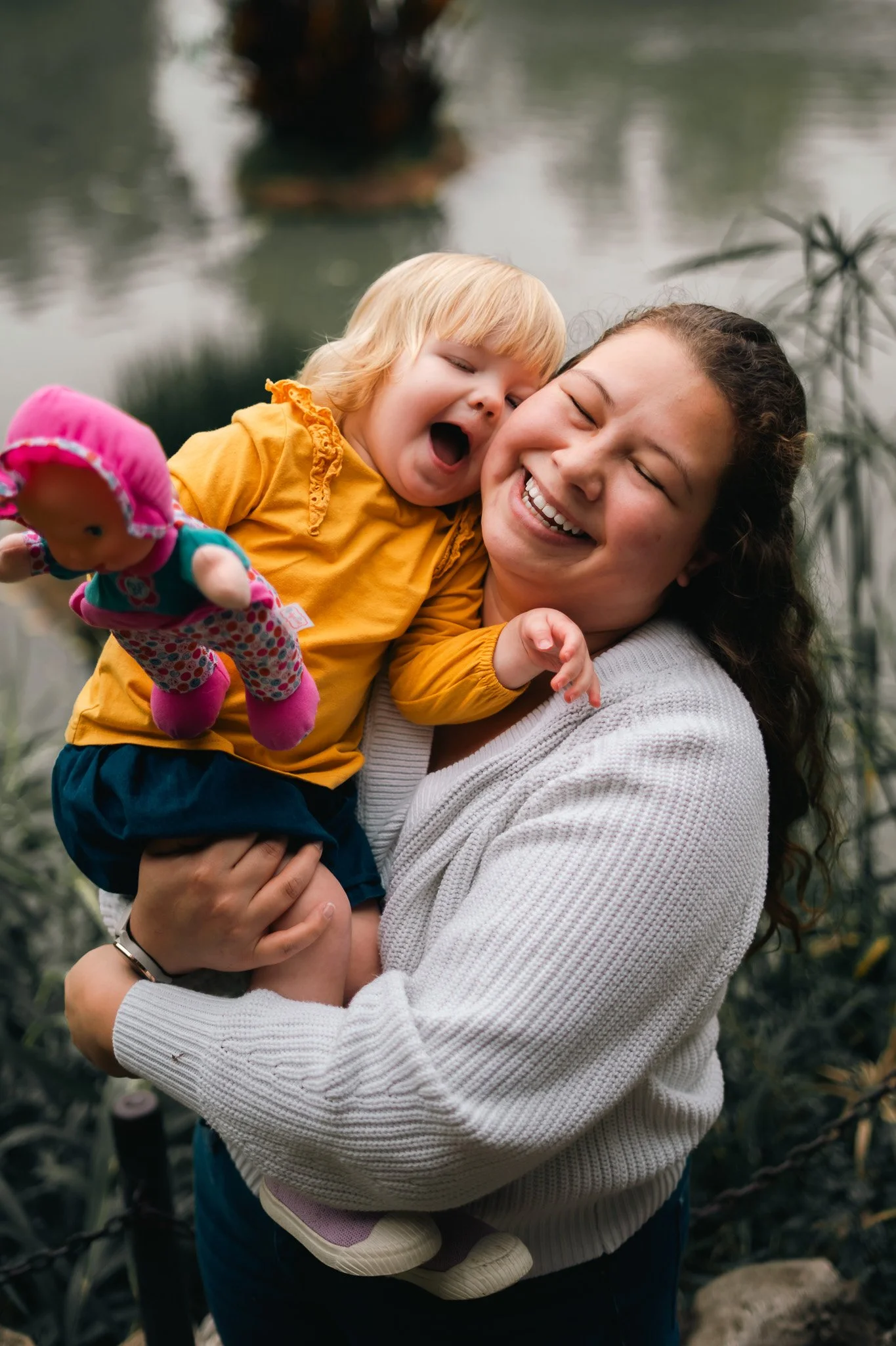 A woman with dark curly hair holding a laughing blonde toddler girl near a body of water outdoors.