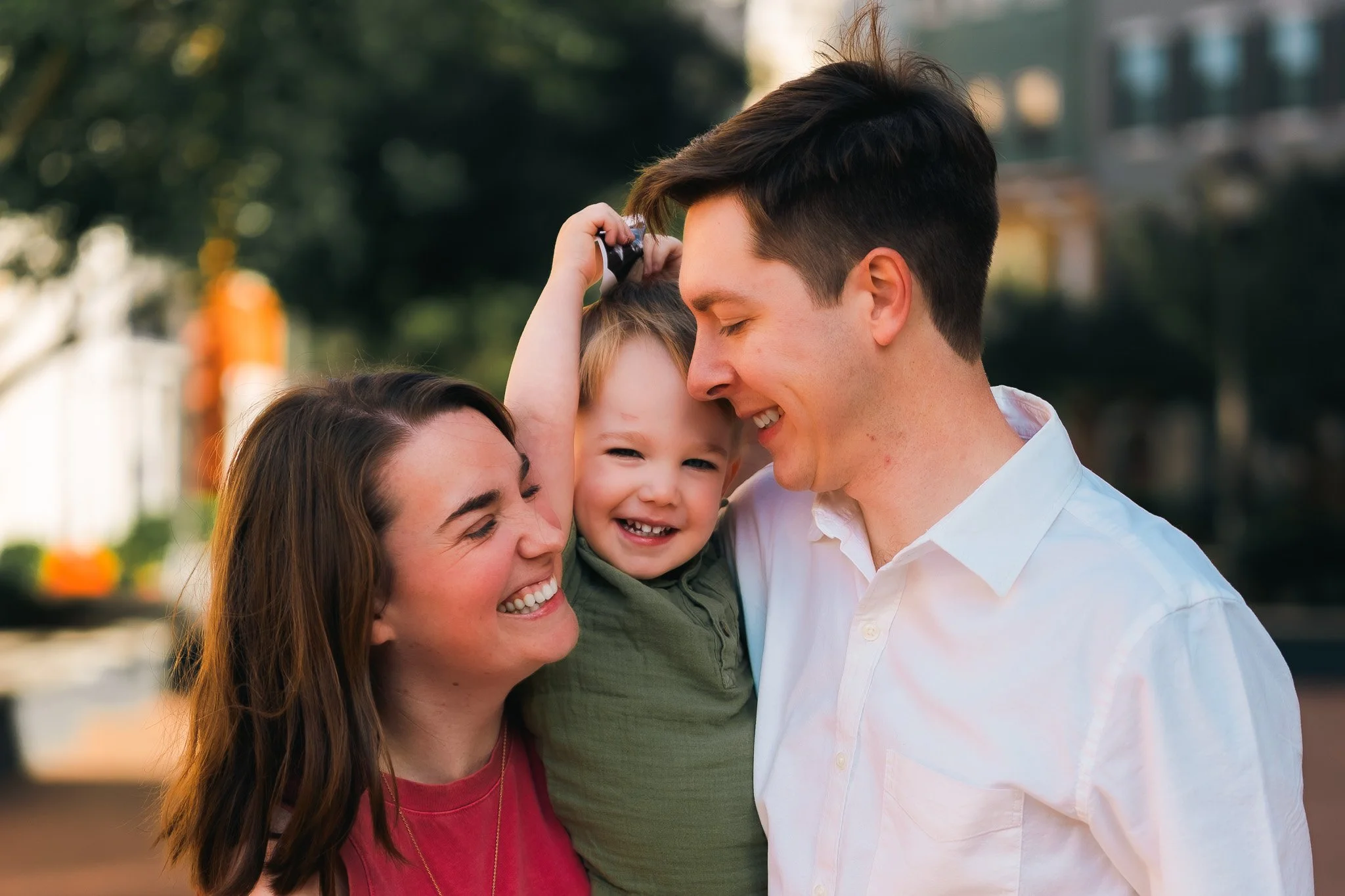 A happy family of three, including a woman, a man, and a young girl, smiling and enjoying a moment together outdoors during the daytime.