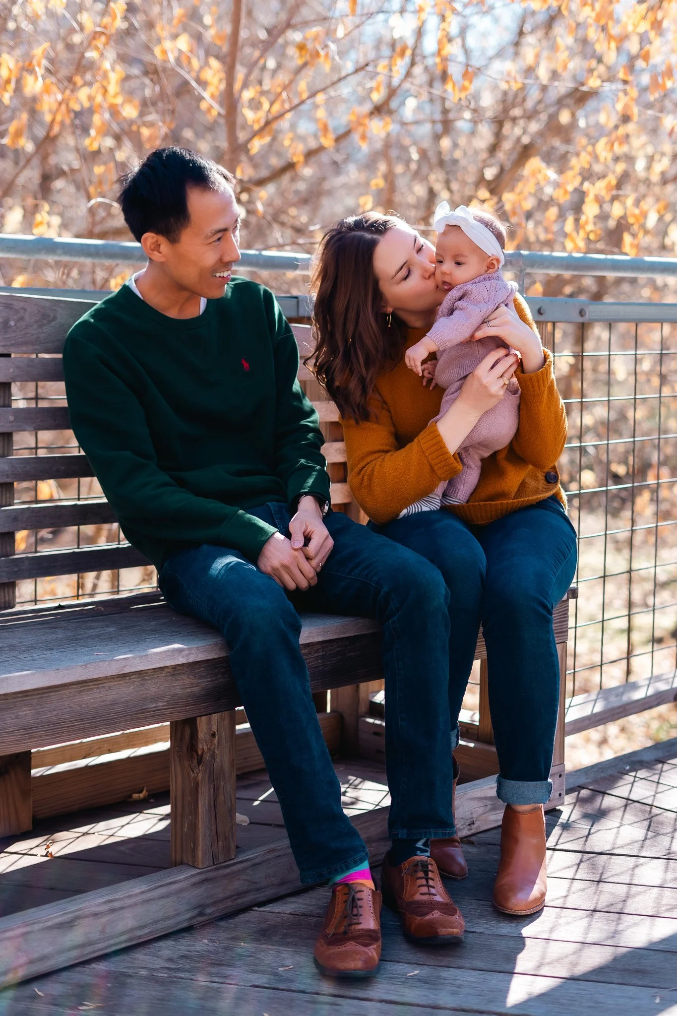 A family of three sitting on an outdoor wooden bench during autumn. The woman is holding and kissing a baby girl, while the man looks on smiling.