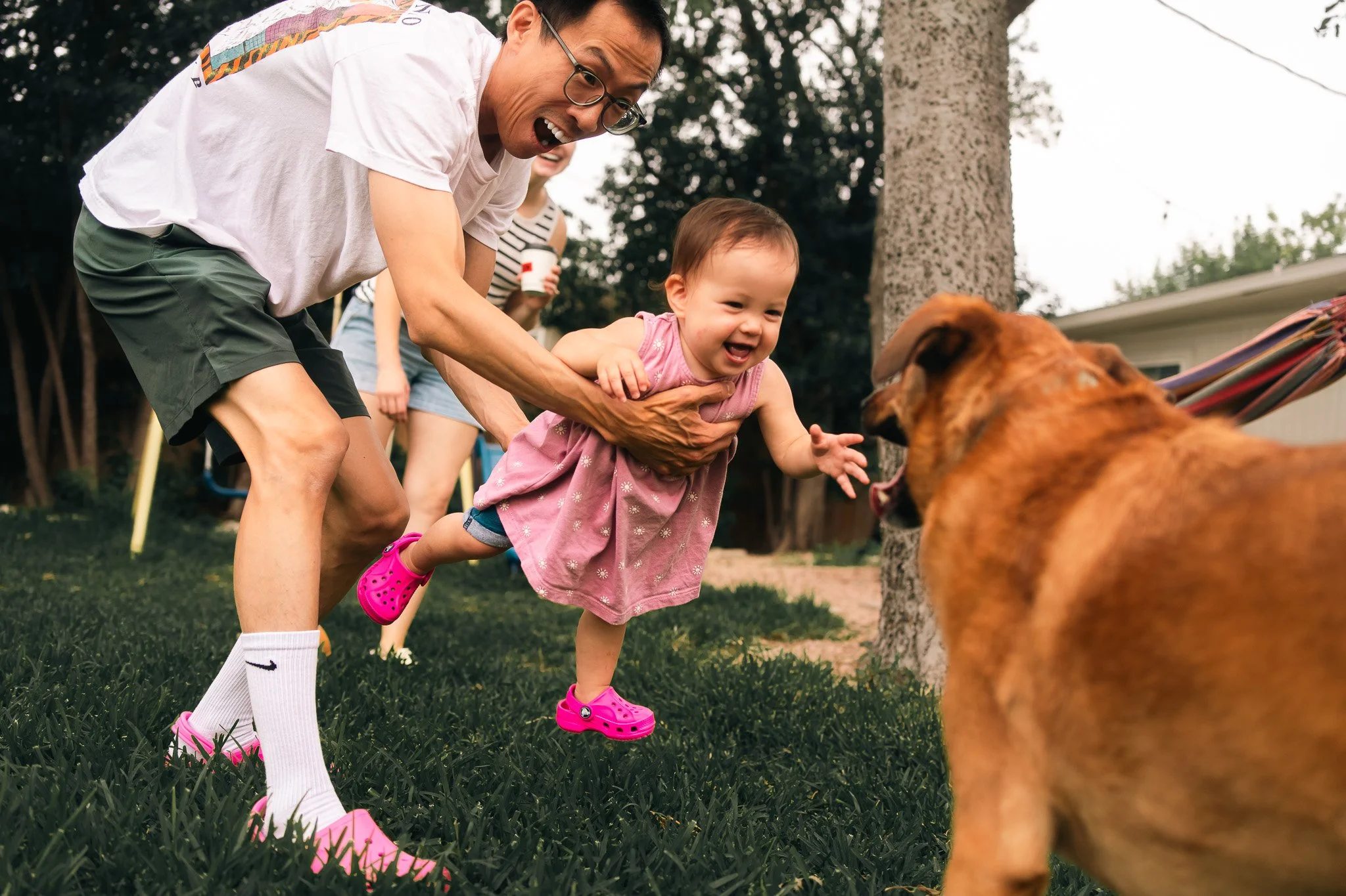 A man and a young girl playing with a dog in a backyard. The man is holding the girl by the waist as she reaches out towards the dog, both smiling and appearing excited. There are trees and a house in the background.