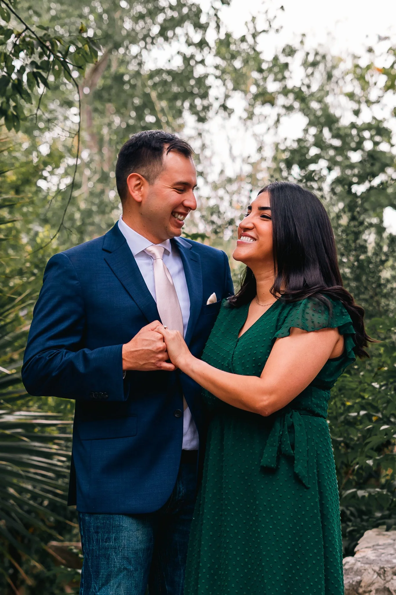 A couple holding hands and smiling at each other outdoors, surrounded by trees and greenery.