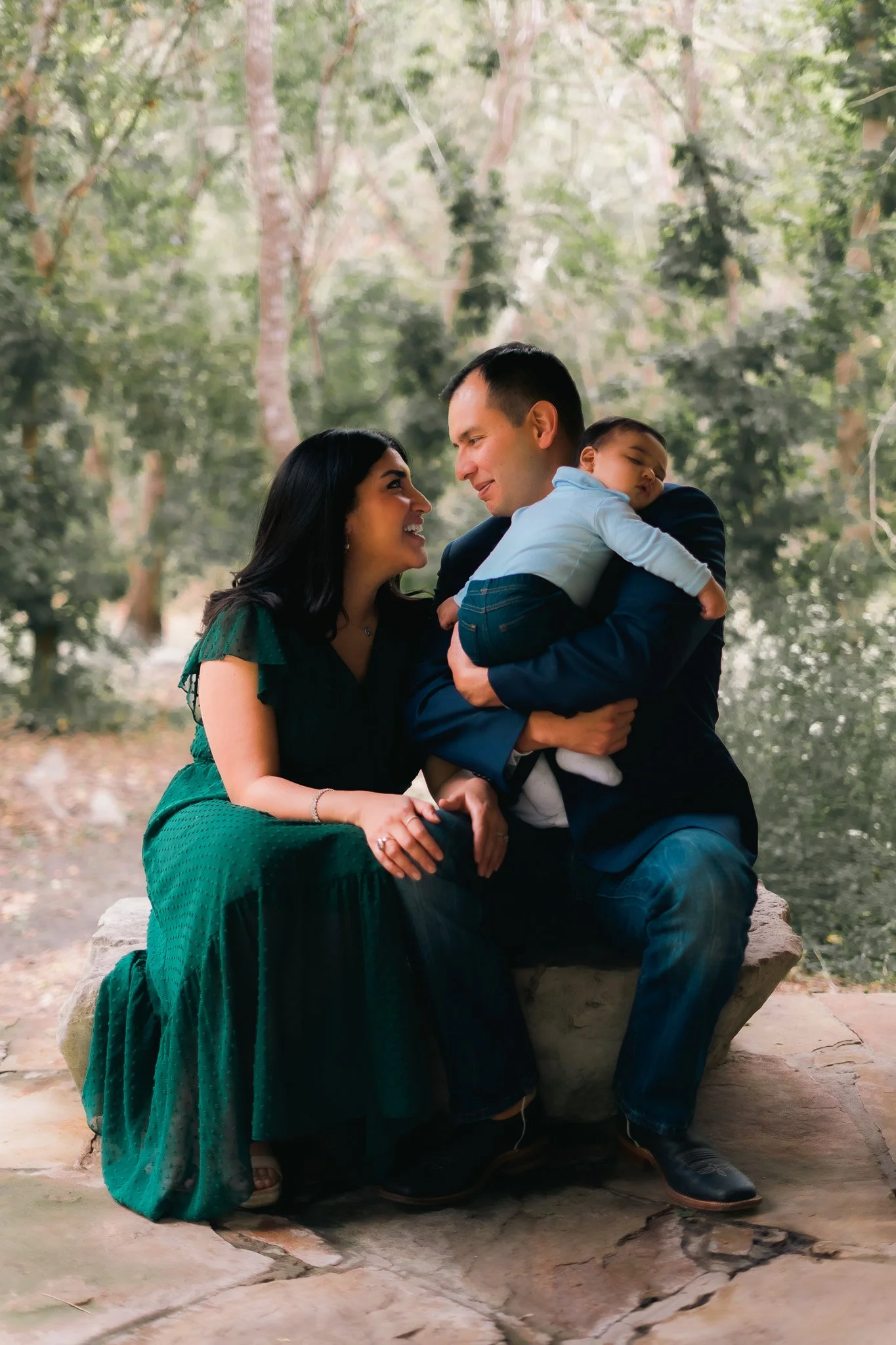 A family sitting outdoors on a stone bench in a wooded area, with a woman in a dark green dress, a man in a dark blazer, and a sleeping child in a light blue shirt and dark pants, engaging in a moment of affection and connection.